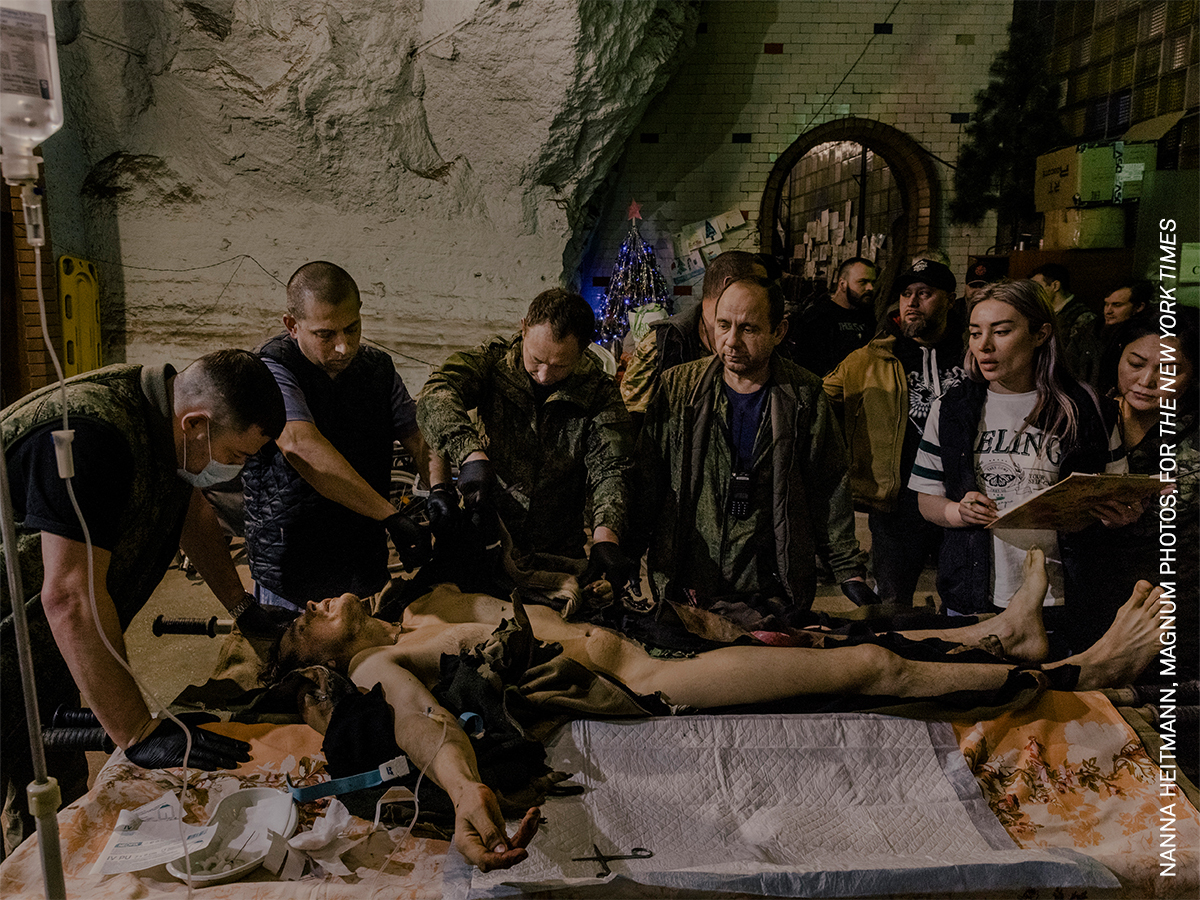 Group of people attending to an injured person lying on a table in a dimly lit room with stone walls.