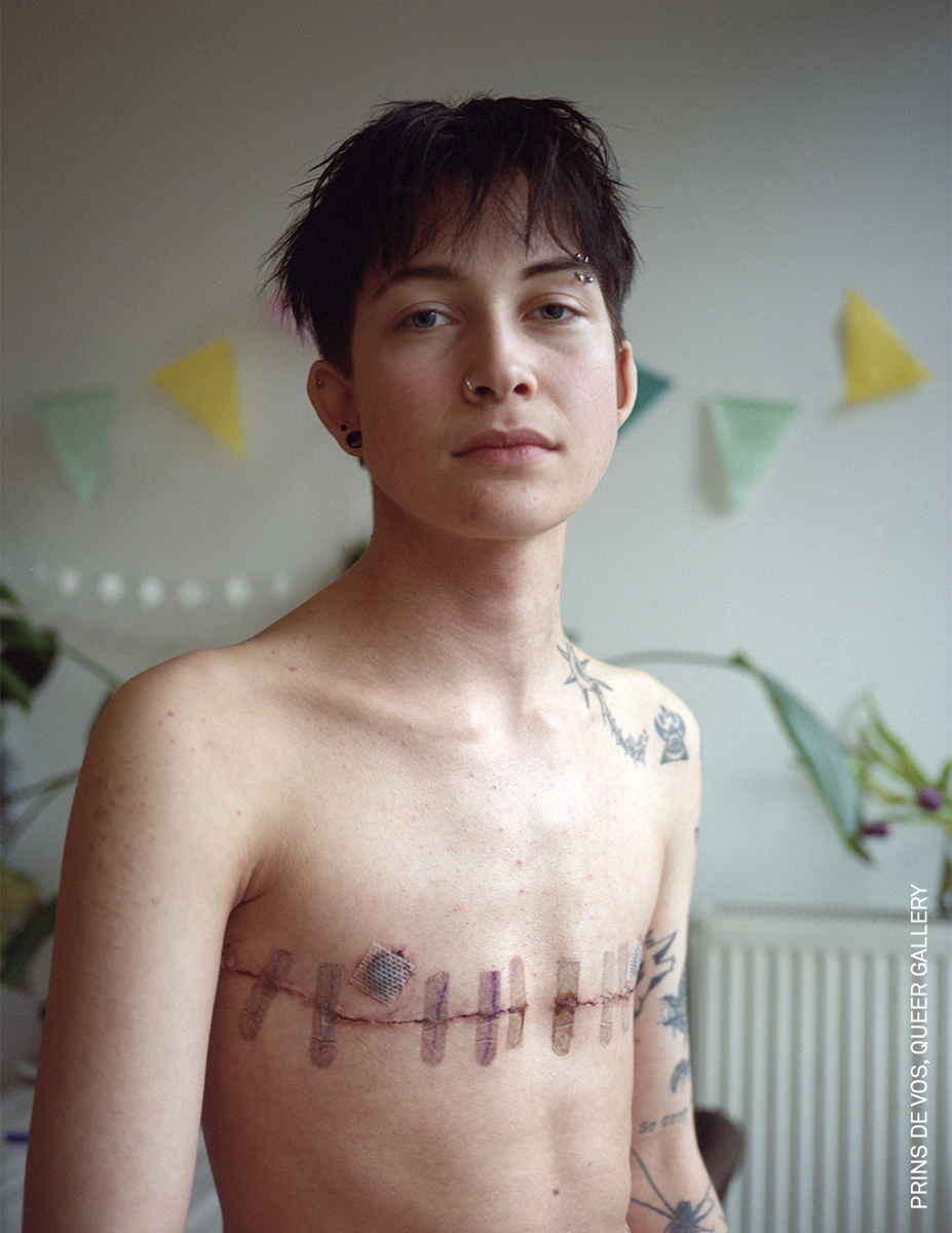 A person with short, dark hair and tattoos on their torso, standing in a room with colourful decorations.