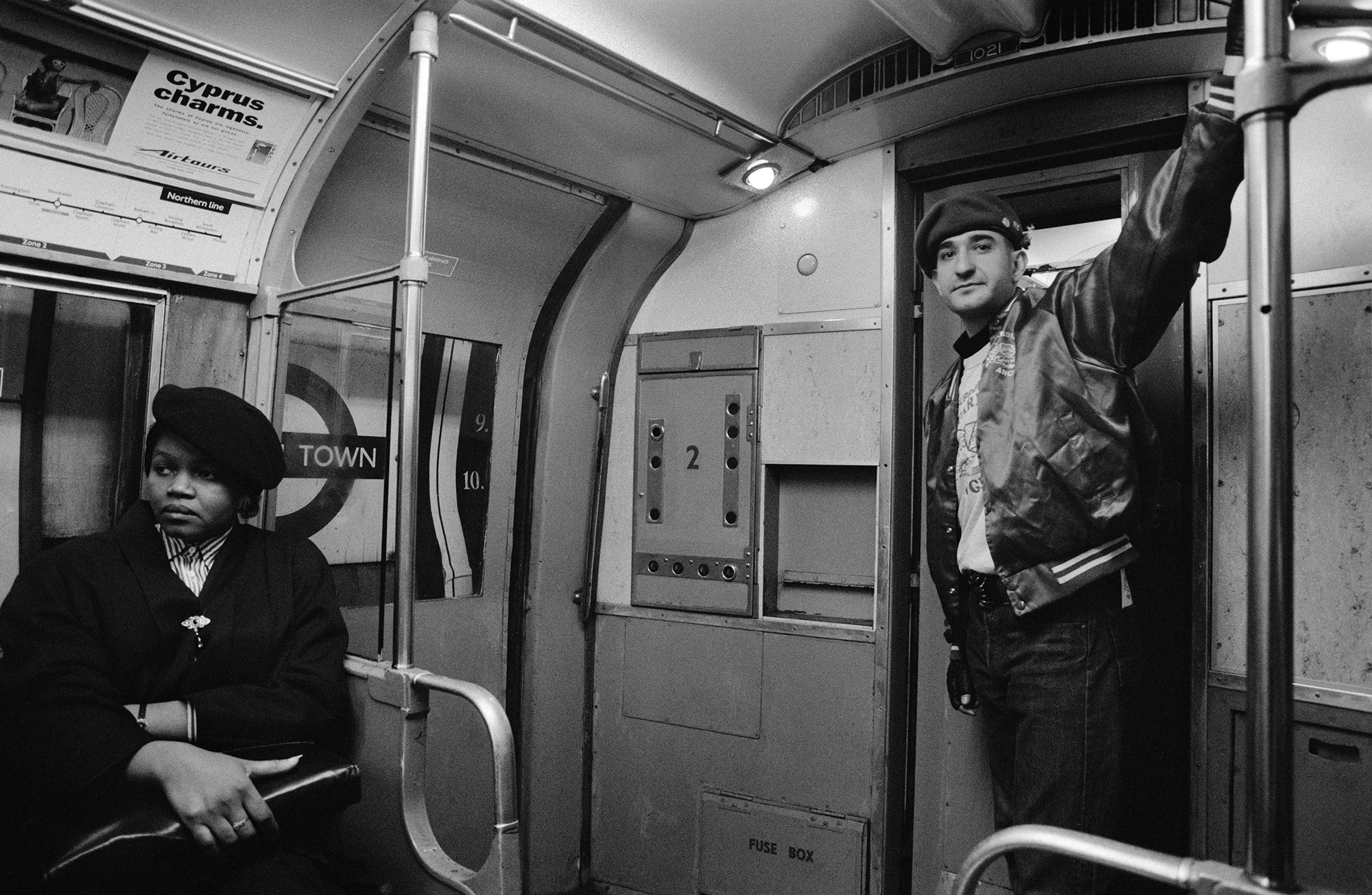 Black and white Underground train carriage interior with seated passenger in dark coat and standing man in cap holding handrail.
