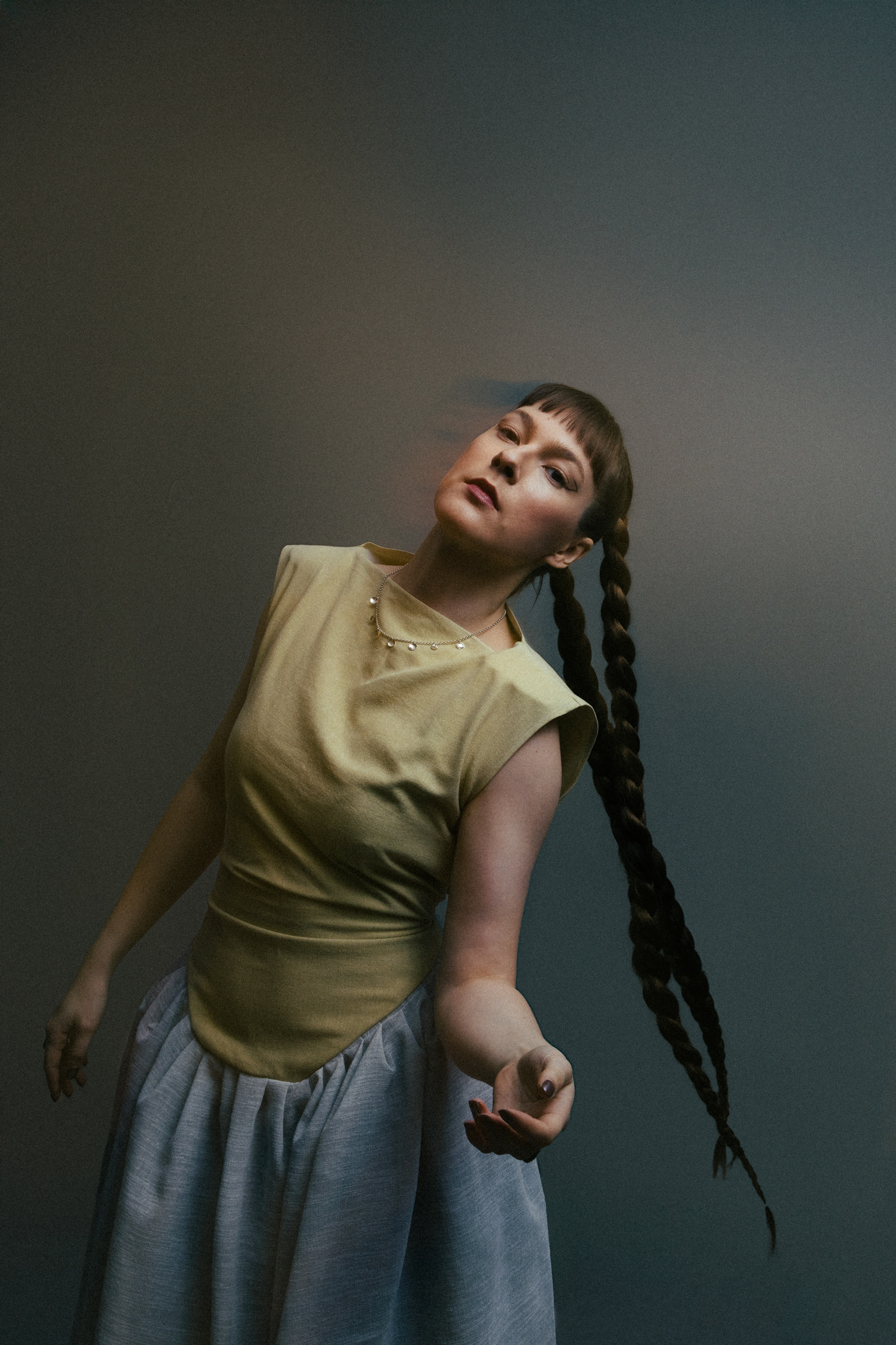 Person with long braided hair wearing olive green top and blue skirt, head tilted back against gradient green-grey background.