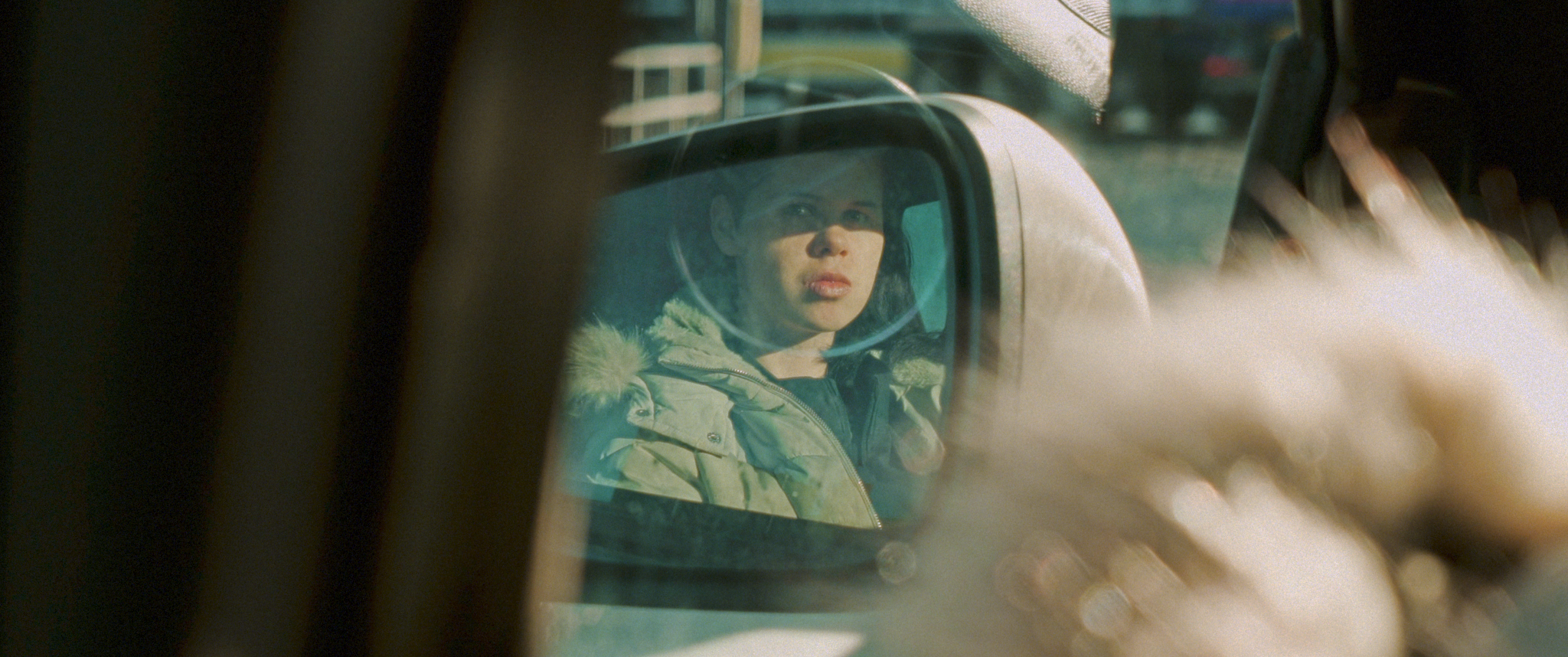 A person in a car wearing a green hooded jacket, visible in the car's side mirror.