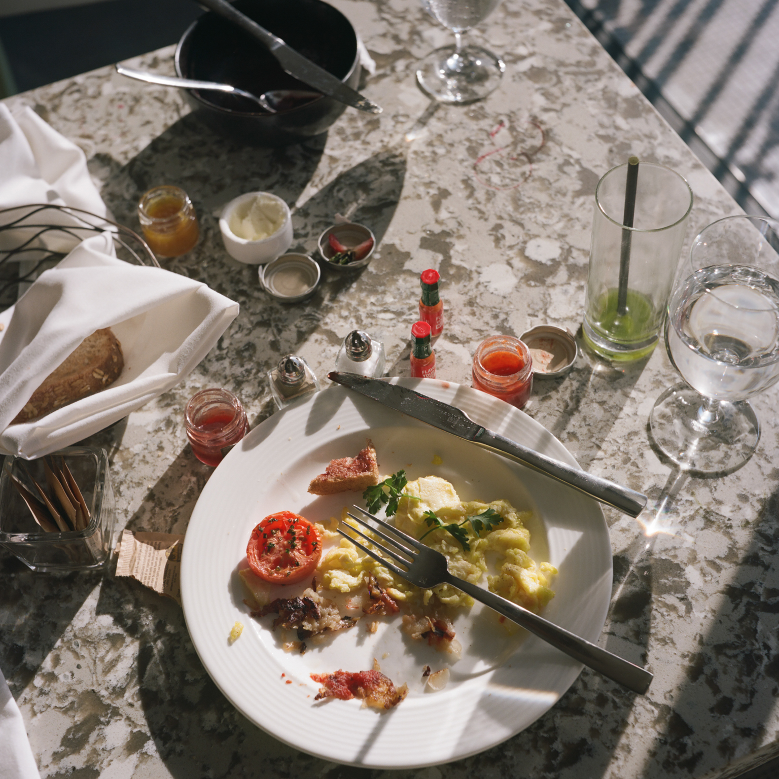 Marble table with white plate containing food remnants, fork, knife. Wine glasses, small containers with coloured liquids, and dining items scattered about.