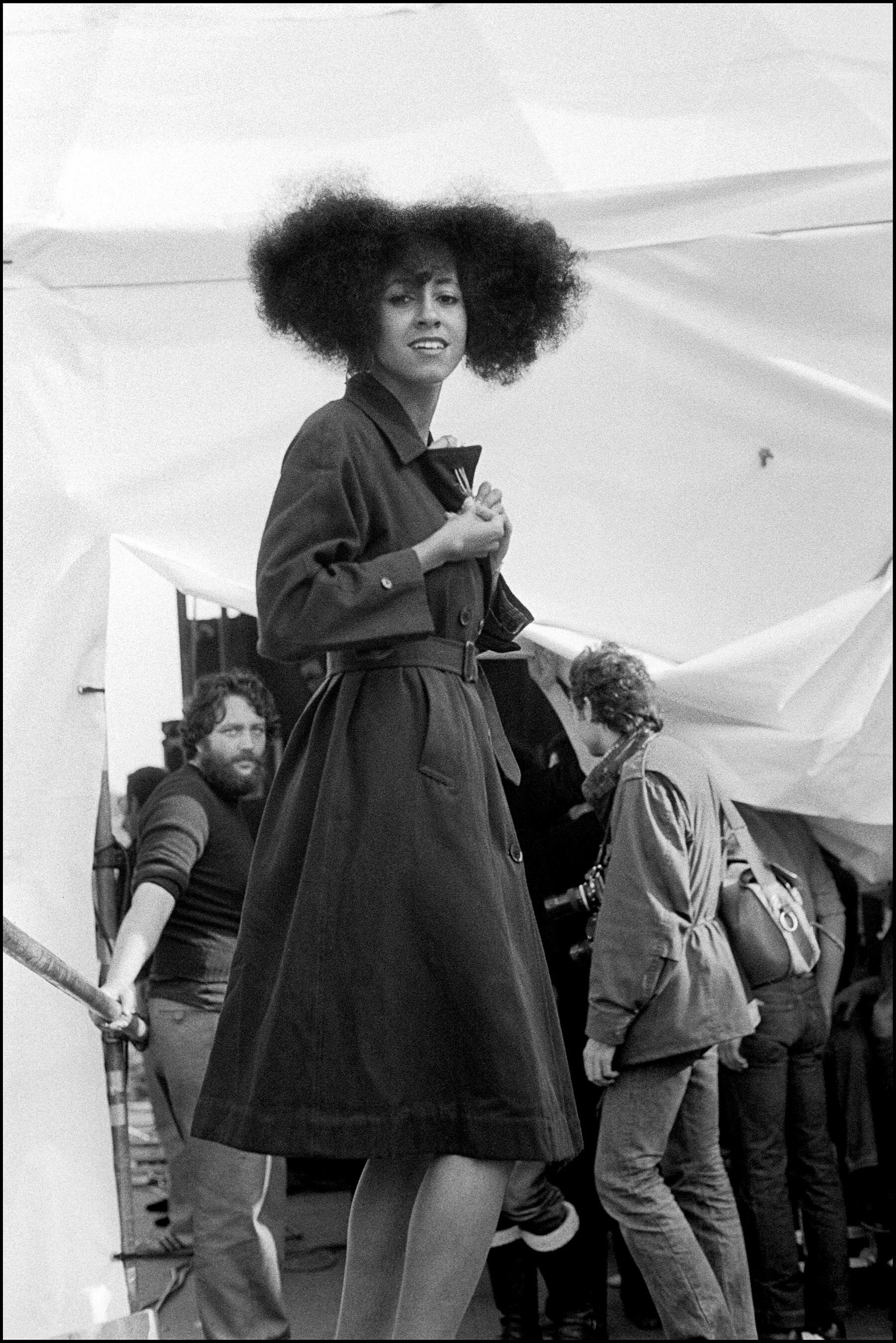 A woman with a large, curly afro hairstyle wearing a long dark coat stands outdoors, with other people visible in the background.