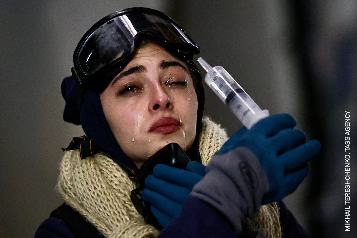 Helmet-wearing woman in cold-weather gear holds a syringe, looking pensive.