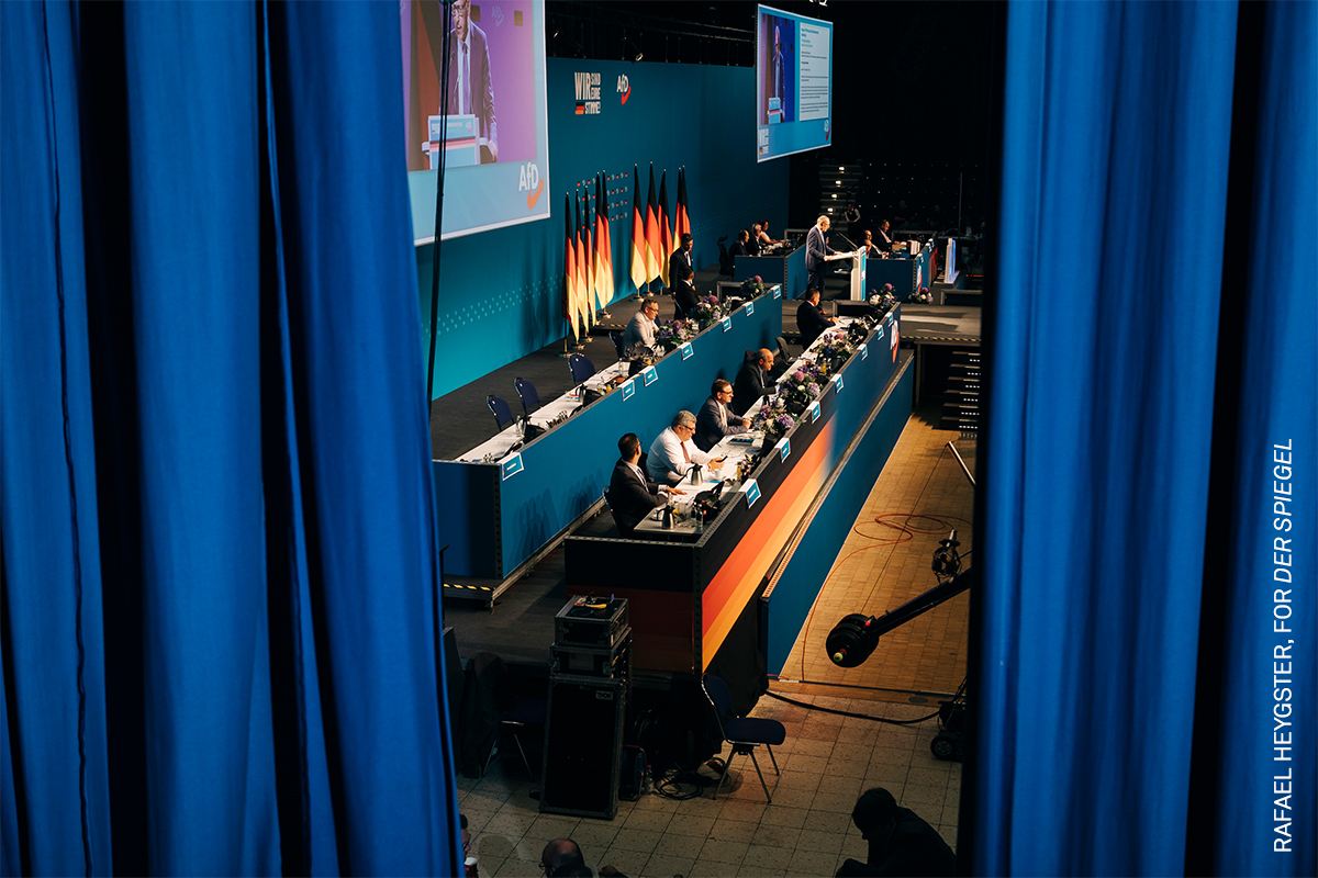Crowded auditorium with rows of seated people, dark blue curtains lining the walls, and lighting equipment visible on the stage.