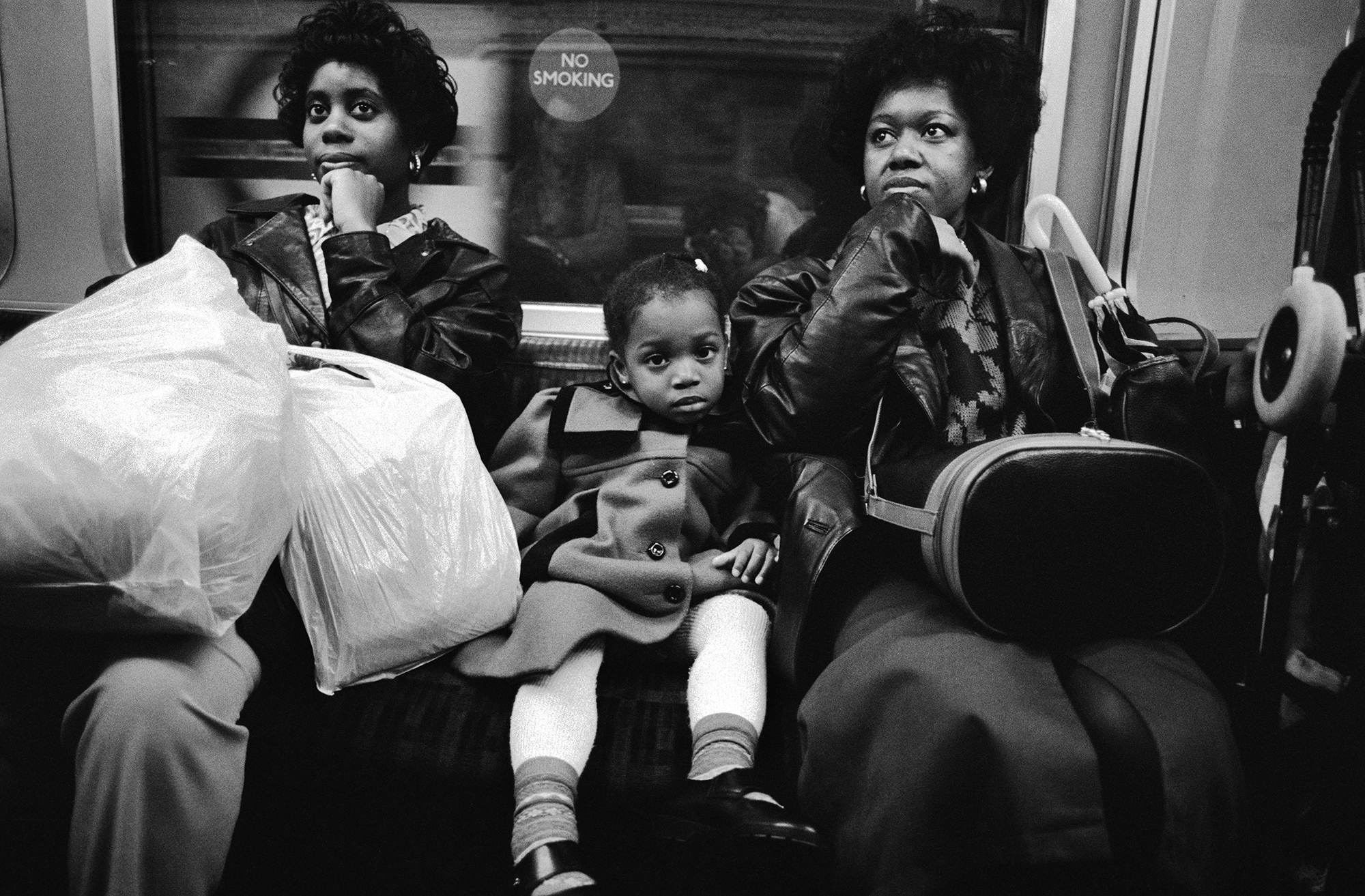 Two women and two children sitting on public transport, black and white image, woman on left holds large white bag.