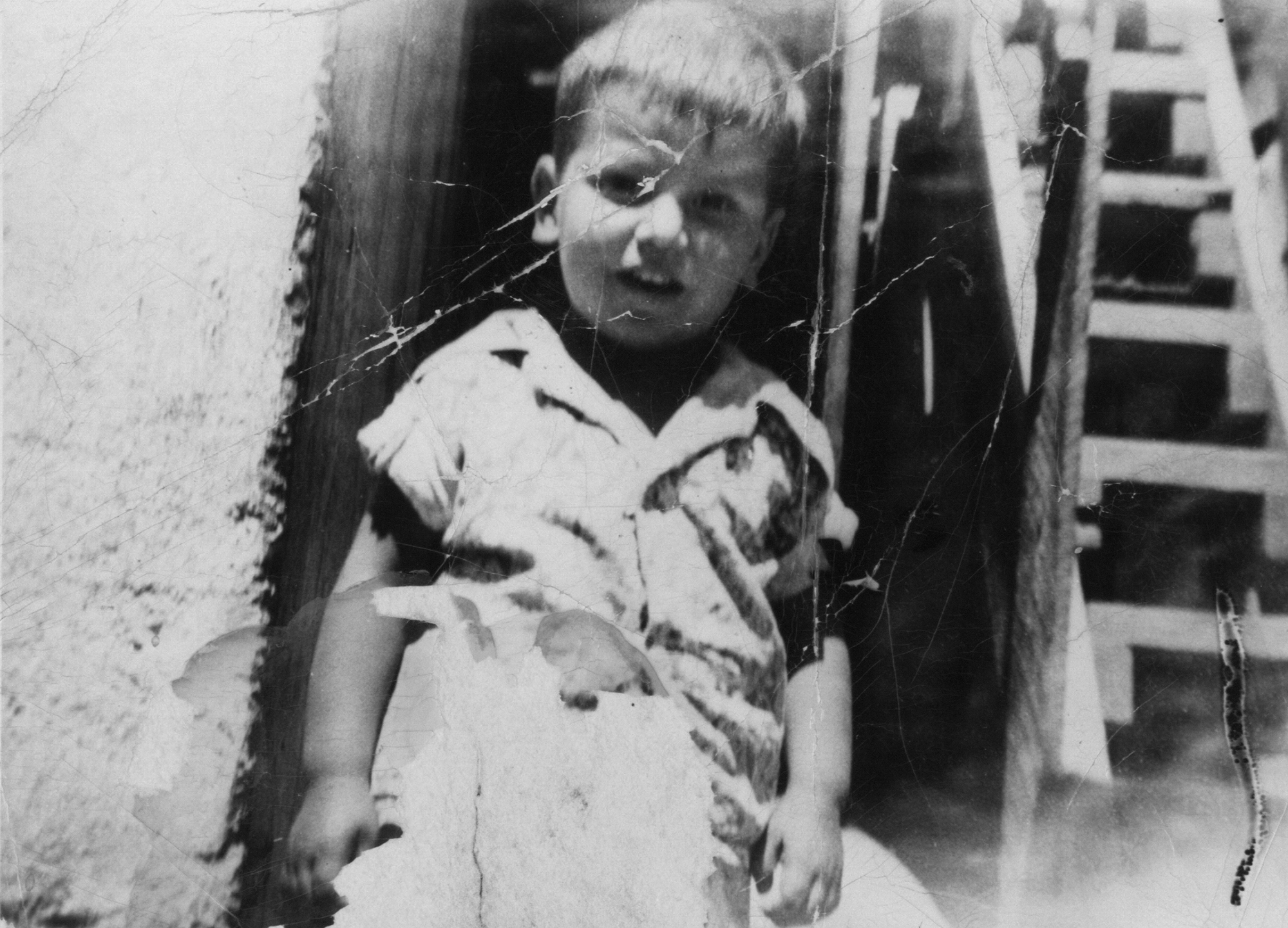 Black and white image of elderly woman in light clothing sitting in wooden chair, with stairs visible in background.