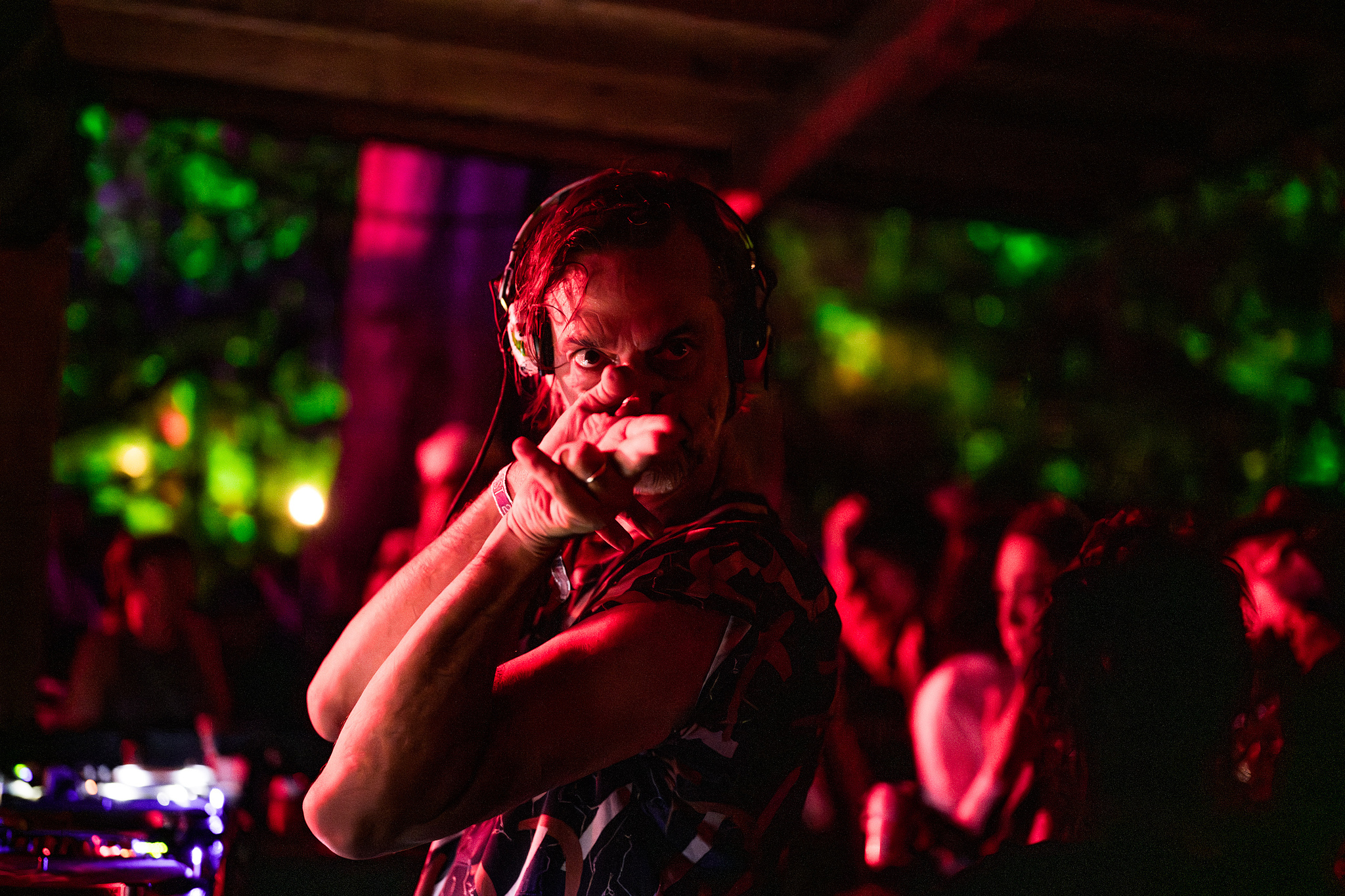 DJ wearing headphones at mixing desk, bathed in red and green stage lighting with blurred coloured lights in background.