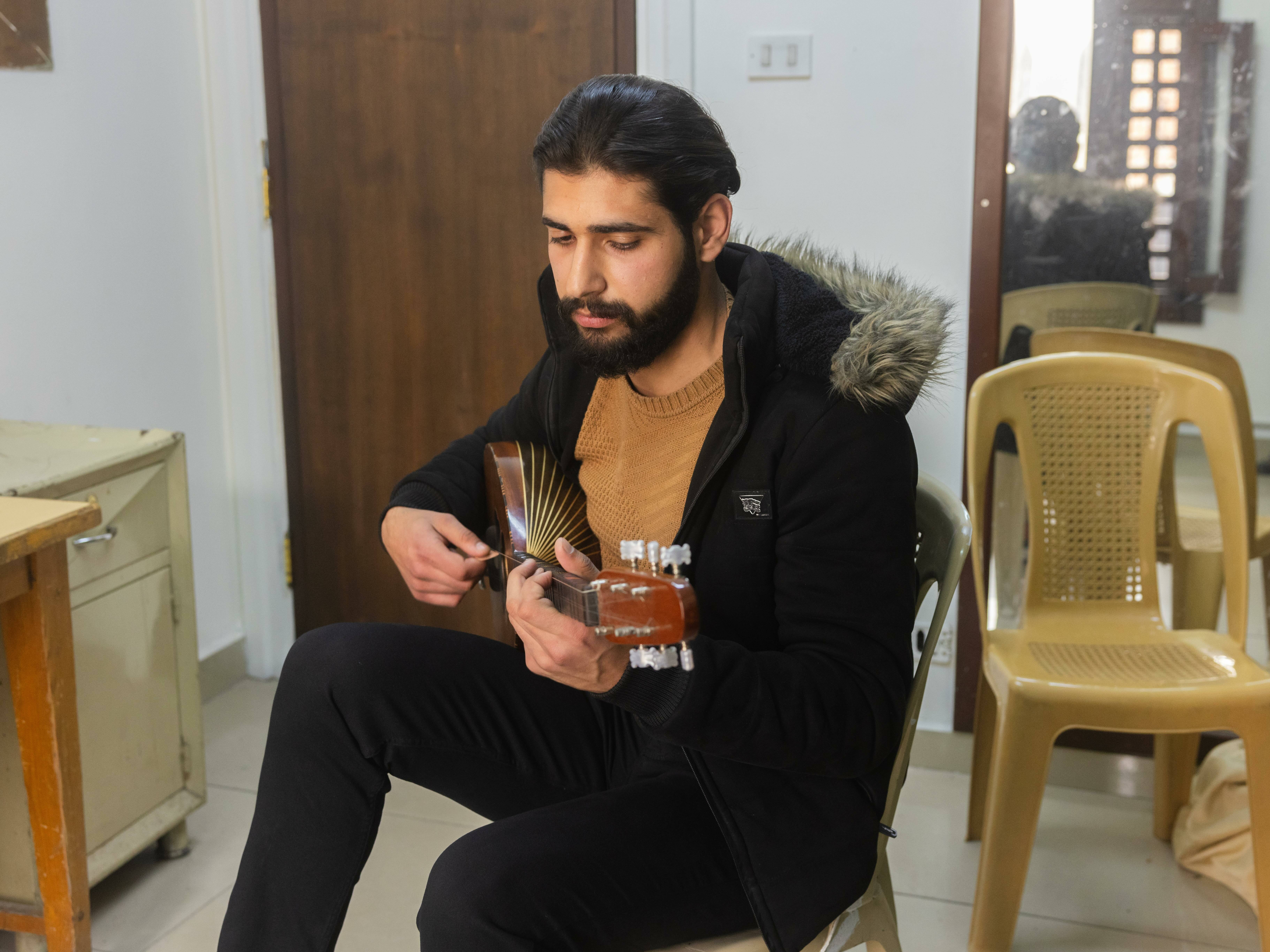 A man with a beard wearing a black jacket and holding a guitar, sitting on a chair in an indoor setting.