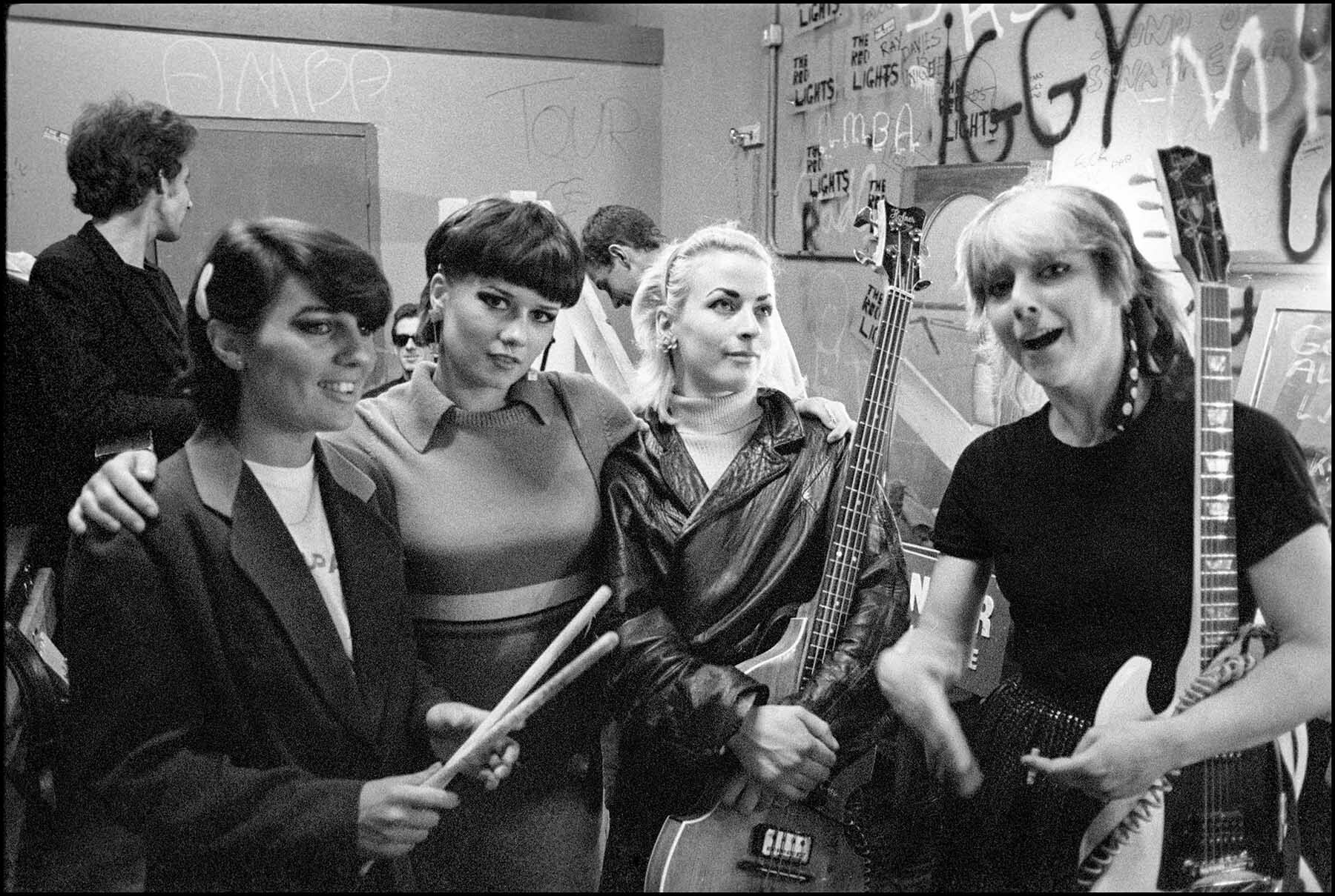 Five individuals, two women and three men, posing together in a room with graffiti-covered walls. The individuals have 1980s-style haircuts and clothing, suggesting the image is from that era. The woman in the centre is holding an electric guitar.