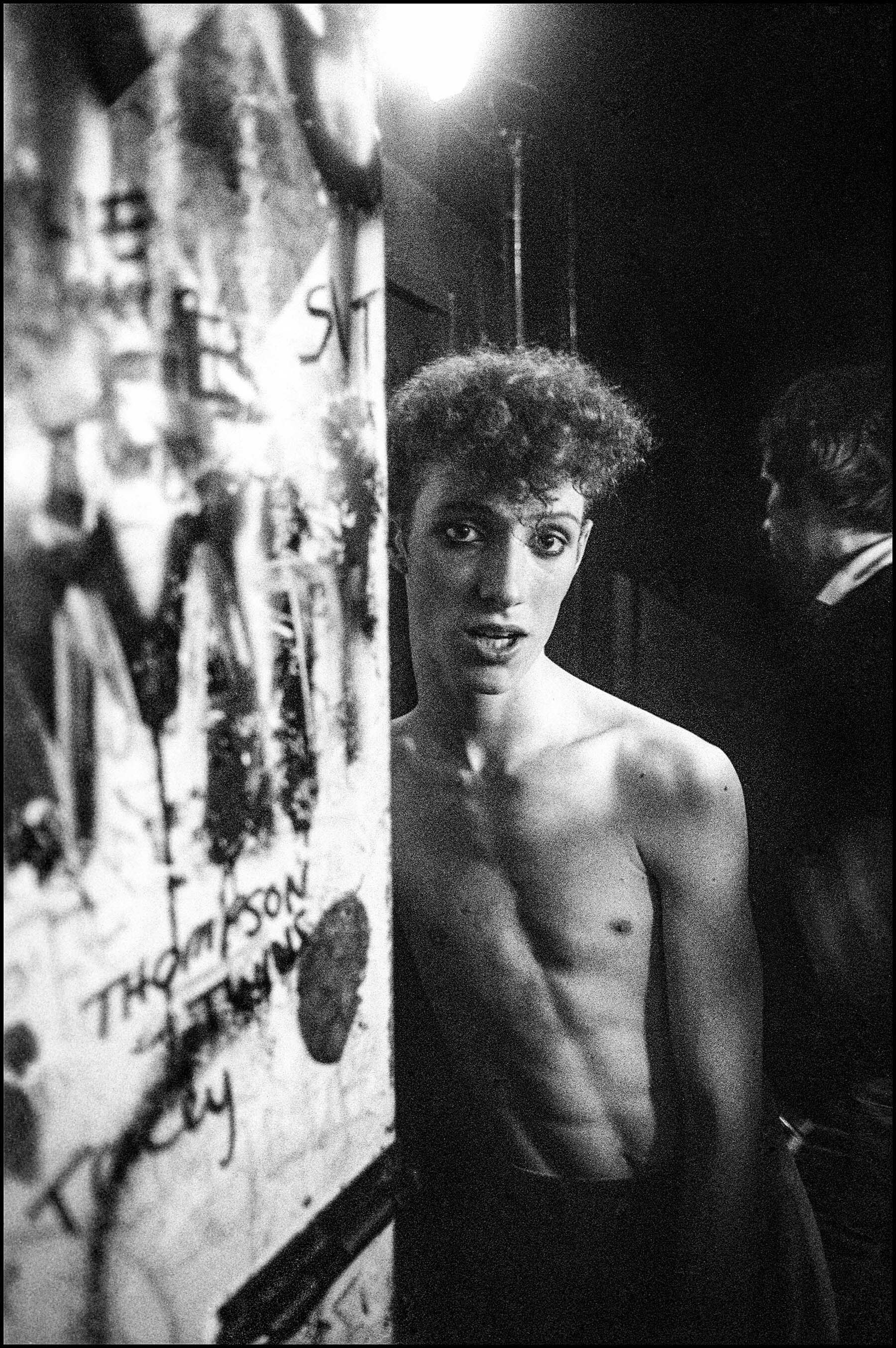 A black-and-white image of a shirtless young man with curly hair standing in front of graffiti-covered walls.