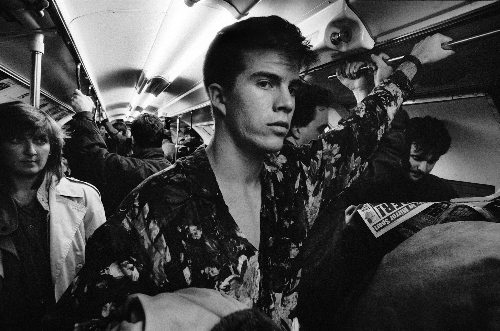 Black and white image of passengers on crowded tube train. Young man in floral shirt sits in foreground, other commuters visible throughout carriage.