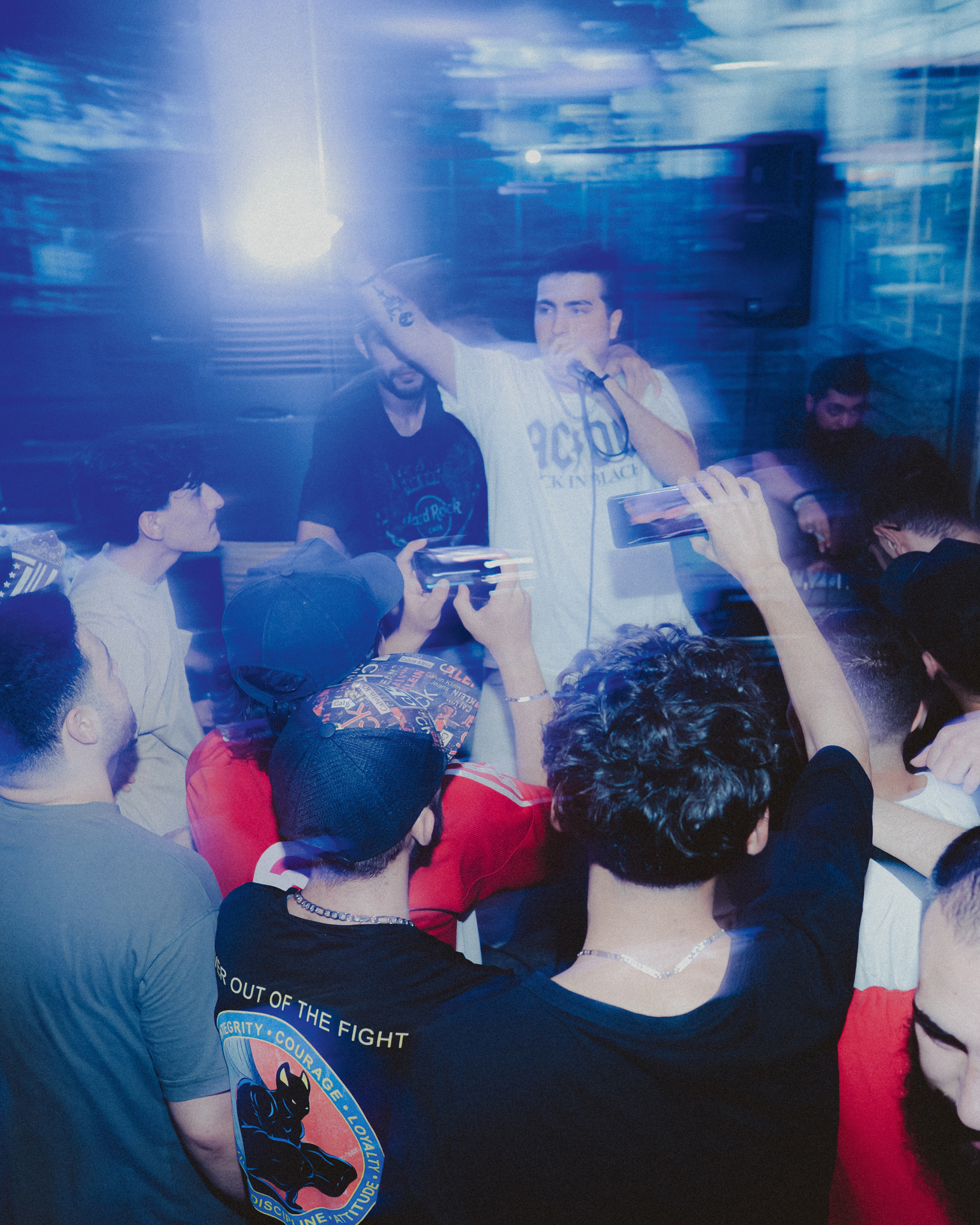 Man in white t-shirt performing on stage with bright overhead lighting, surrounded by audience holding phones, blue-tinted atmosphere.