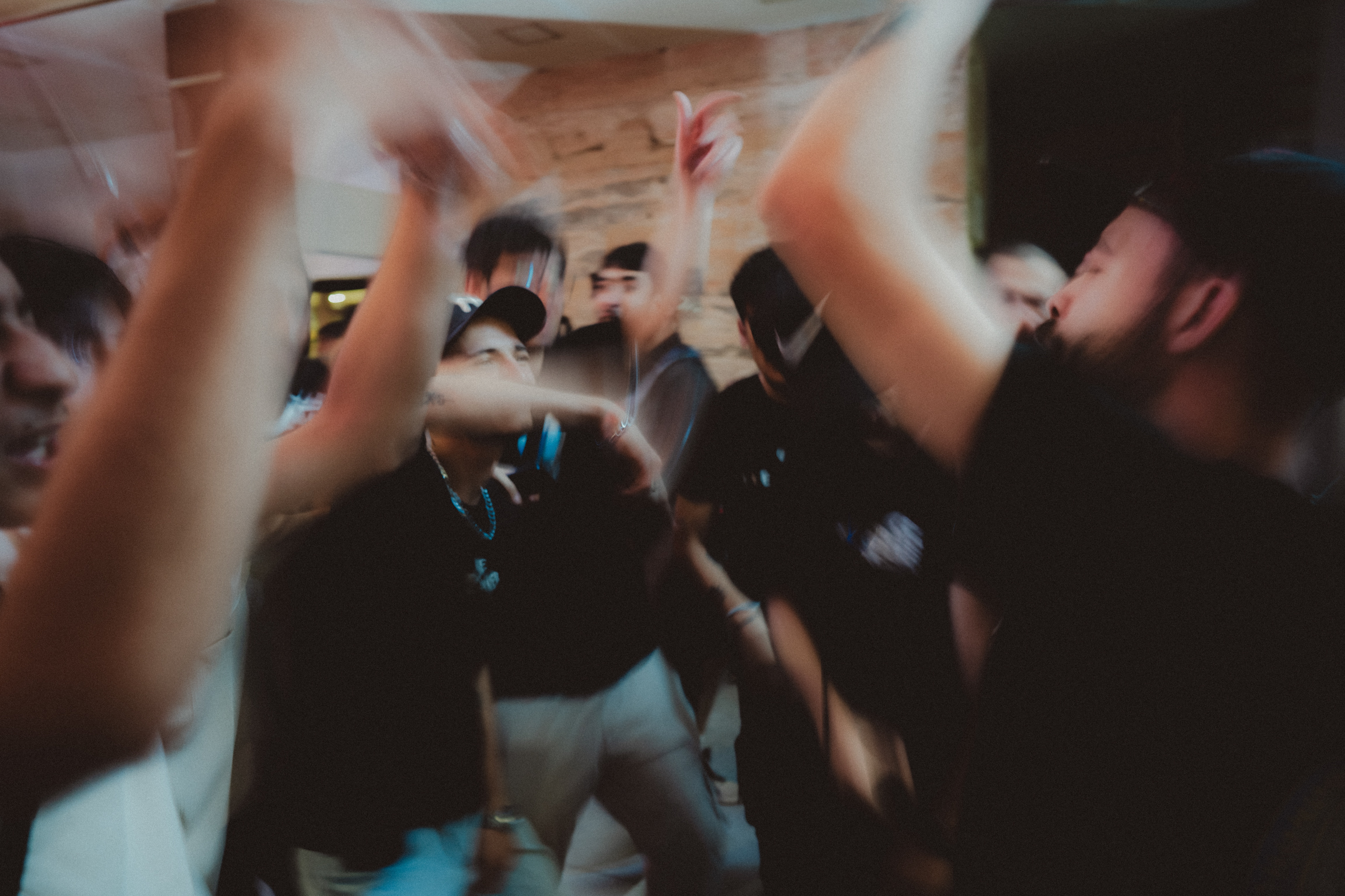 Blurred crowd with raised arms in dimly lit indoor space, motion blur creating streaked effect across the image.