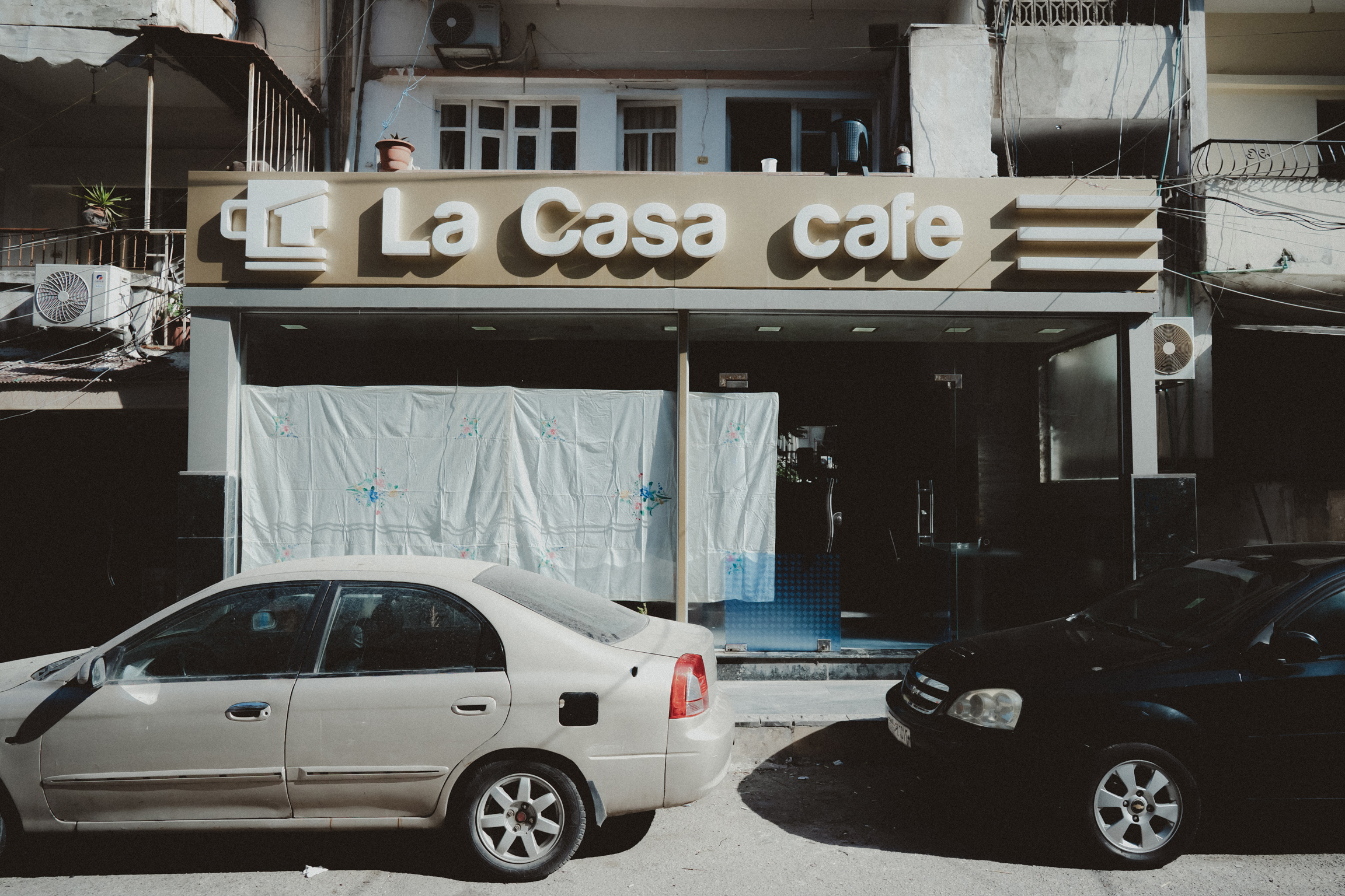Storefront with "Le Casa Cafe" sign above entrance, white plastic sheeting covering doorway, silver and dark cars parked in front.