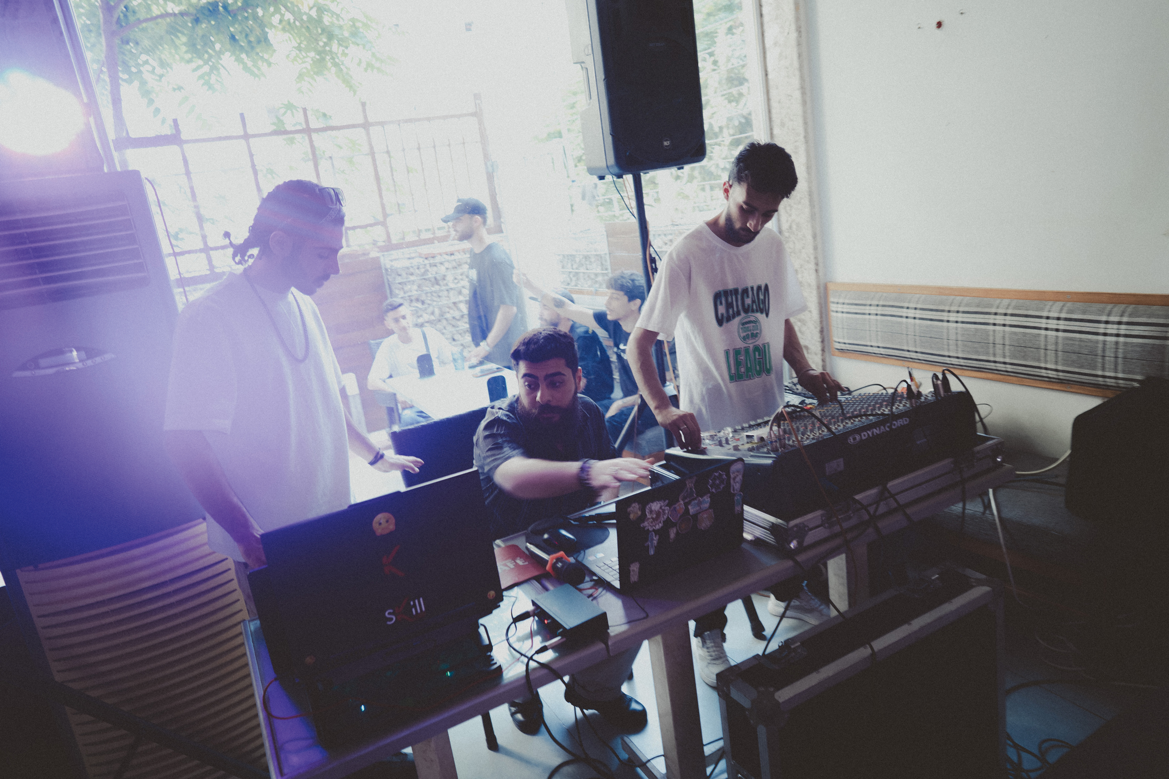 DJ at mixing desk with purple stage lighting, several people gathered around equipment in white-walled room with windows.