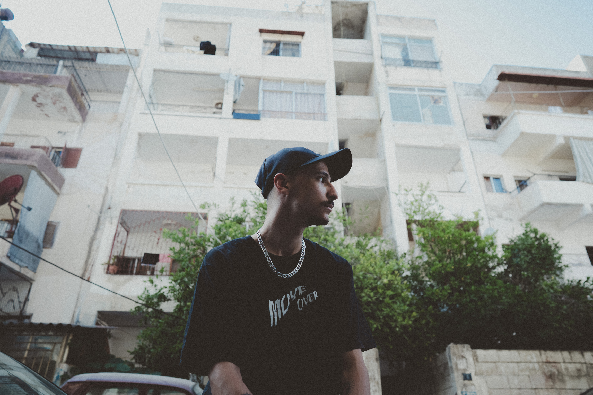 Man in black cap and t-shirt stands in front of white apartment buildings with balconies, green trees visible between structures.