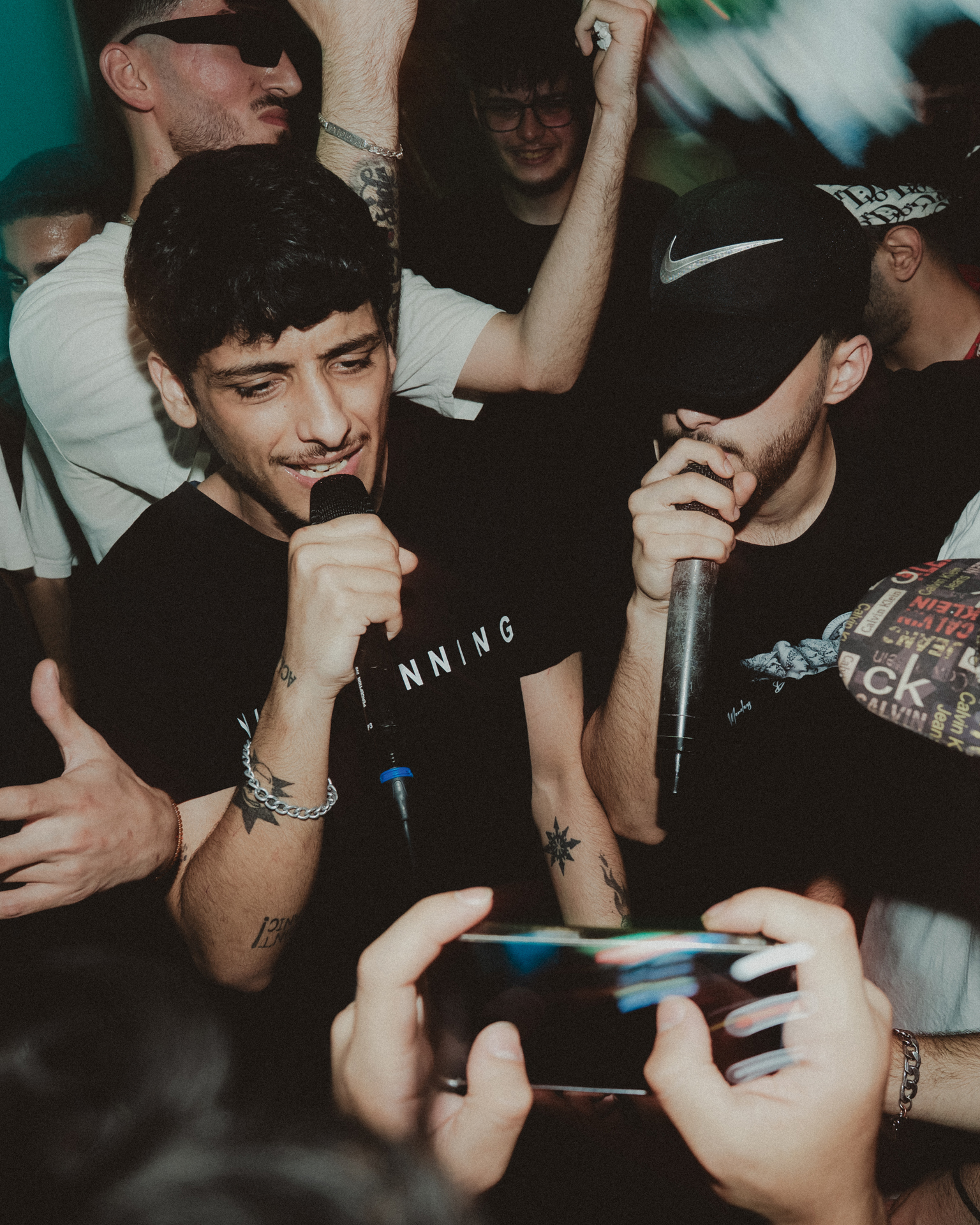 Young man with microphone performing at crowded indoor event, surrounded by audience members with phones recording
