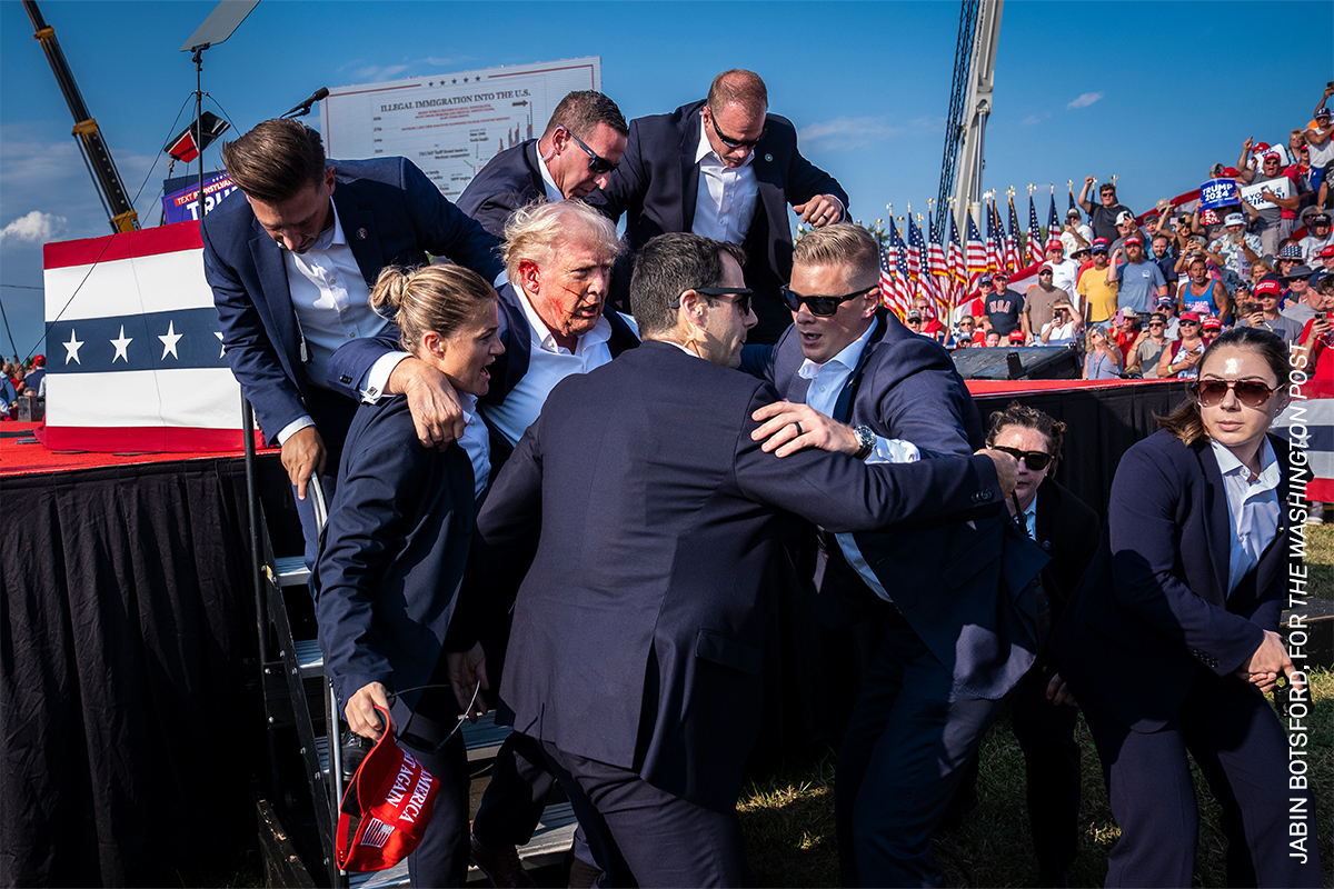 Large crowd of people, mostly men in suits, embracing and interacting with one another in front of an American flag backdrop.