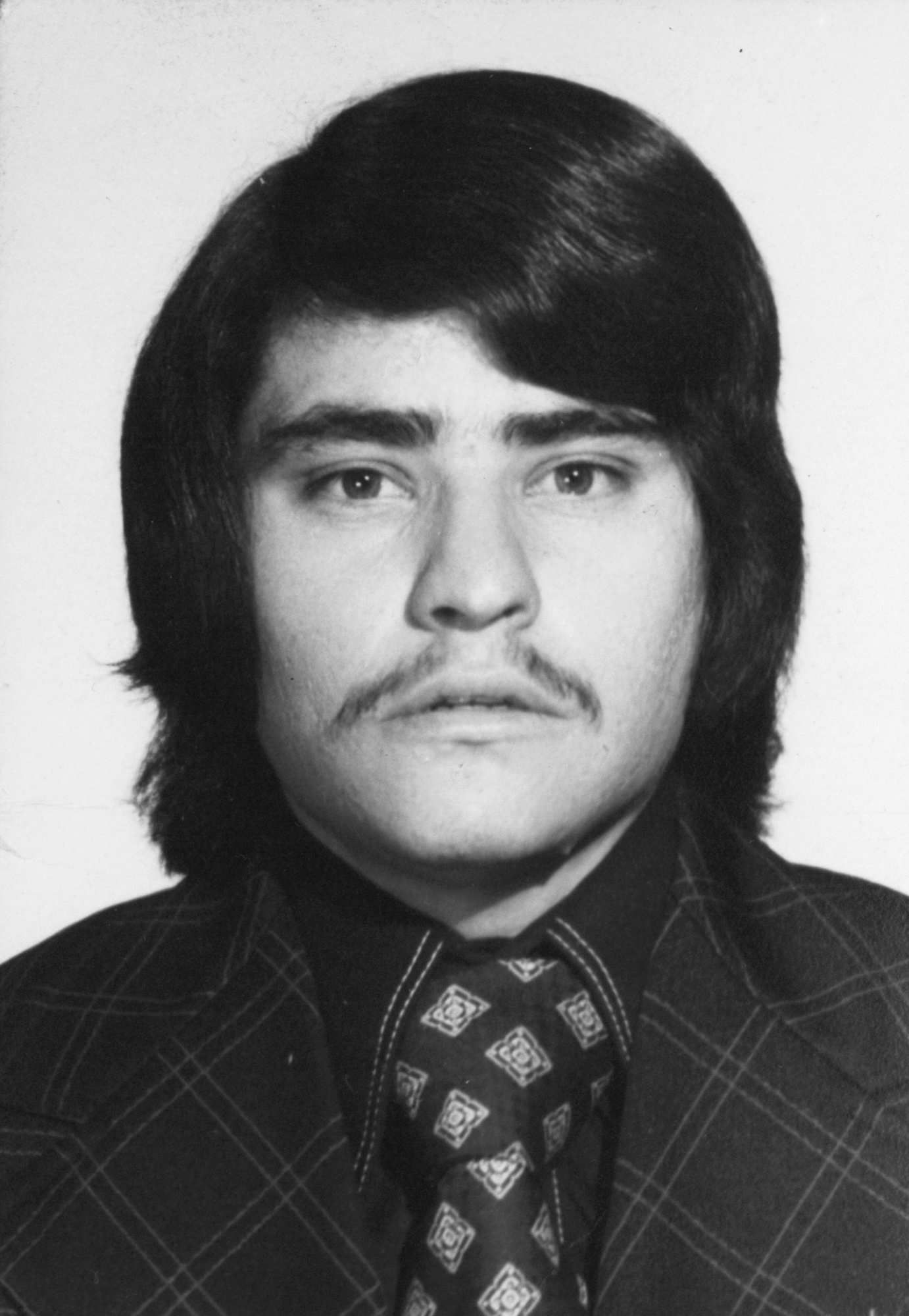 Black and white portrait of young man with dark hair, moustache, wearing patterned tie and suit jacket.