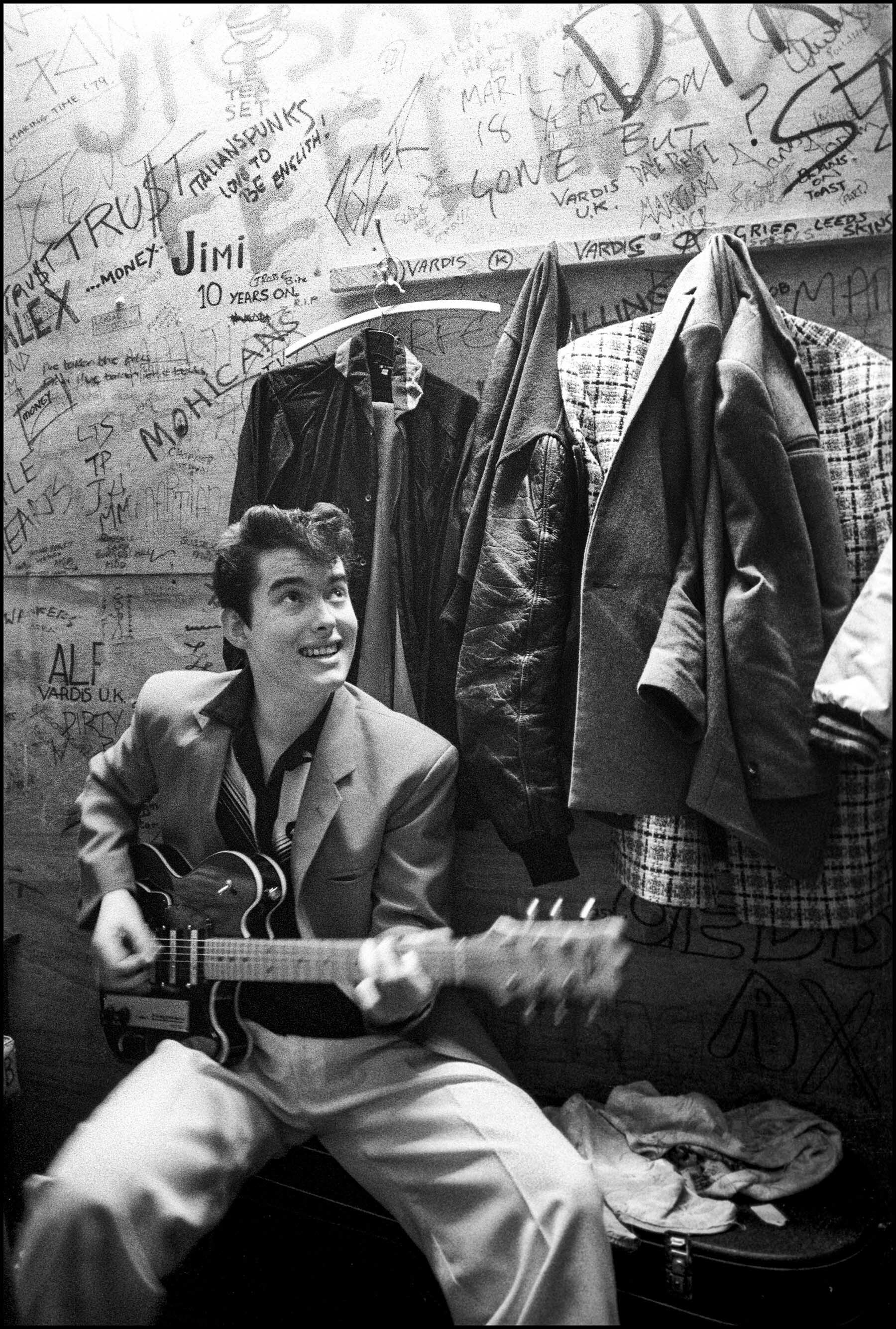 A black and white image of a smiling young man playing an electric guitar in a cluttered room. The background includes graffiti-covered walls, hanging clothes, and various items scattered around.