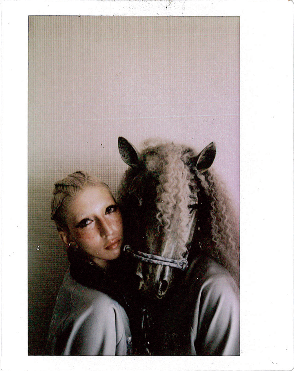 Monochrome image of a person with curly hair and horse-like ears posing with a stuffed animal resembling a horse.
