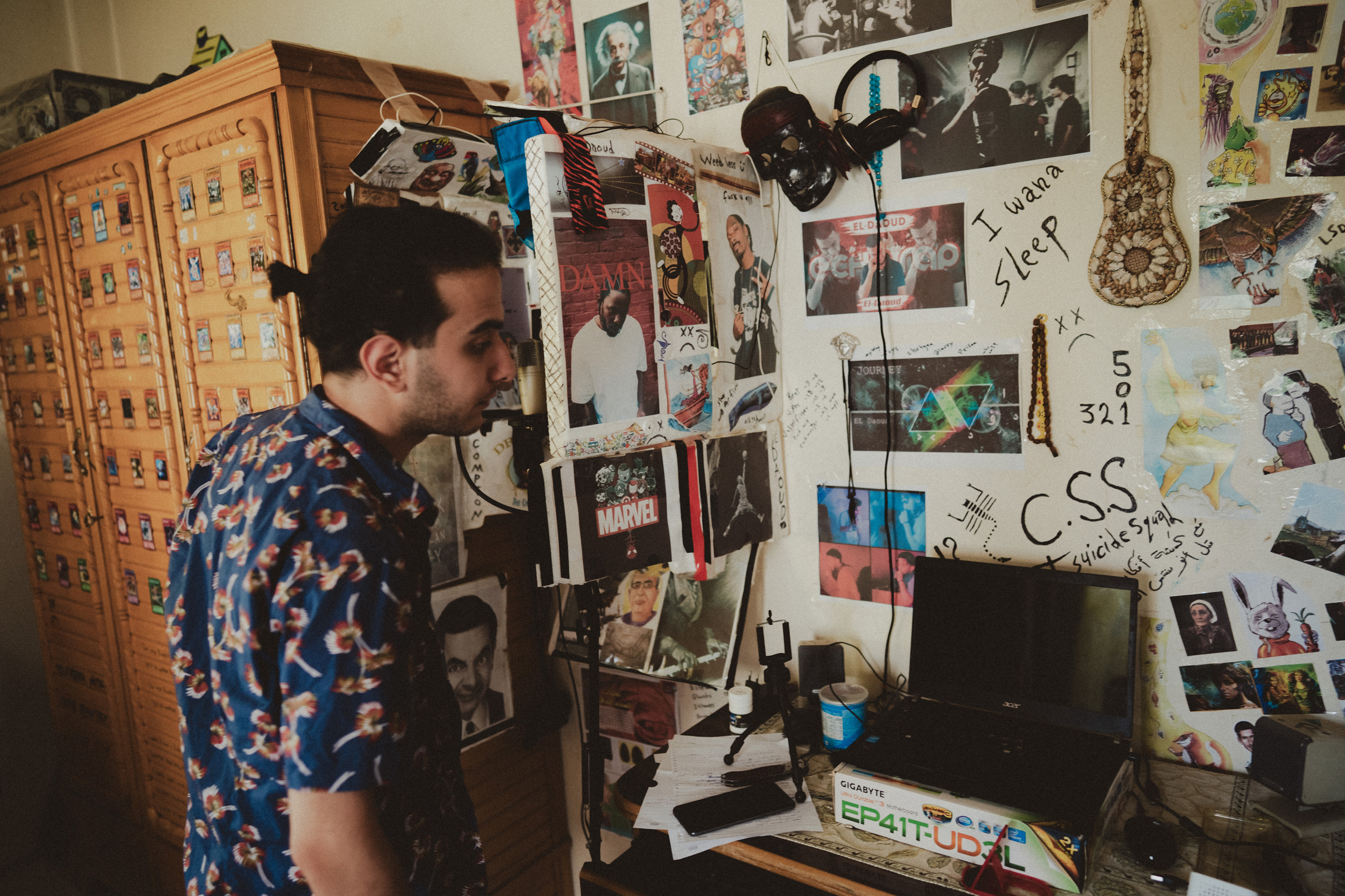 Man in patterned shirt sits at desk with computer in room covered with photos, posters, and memorabilia on walls and shelves.