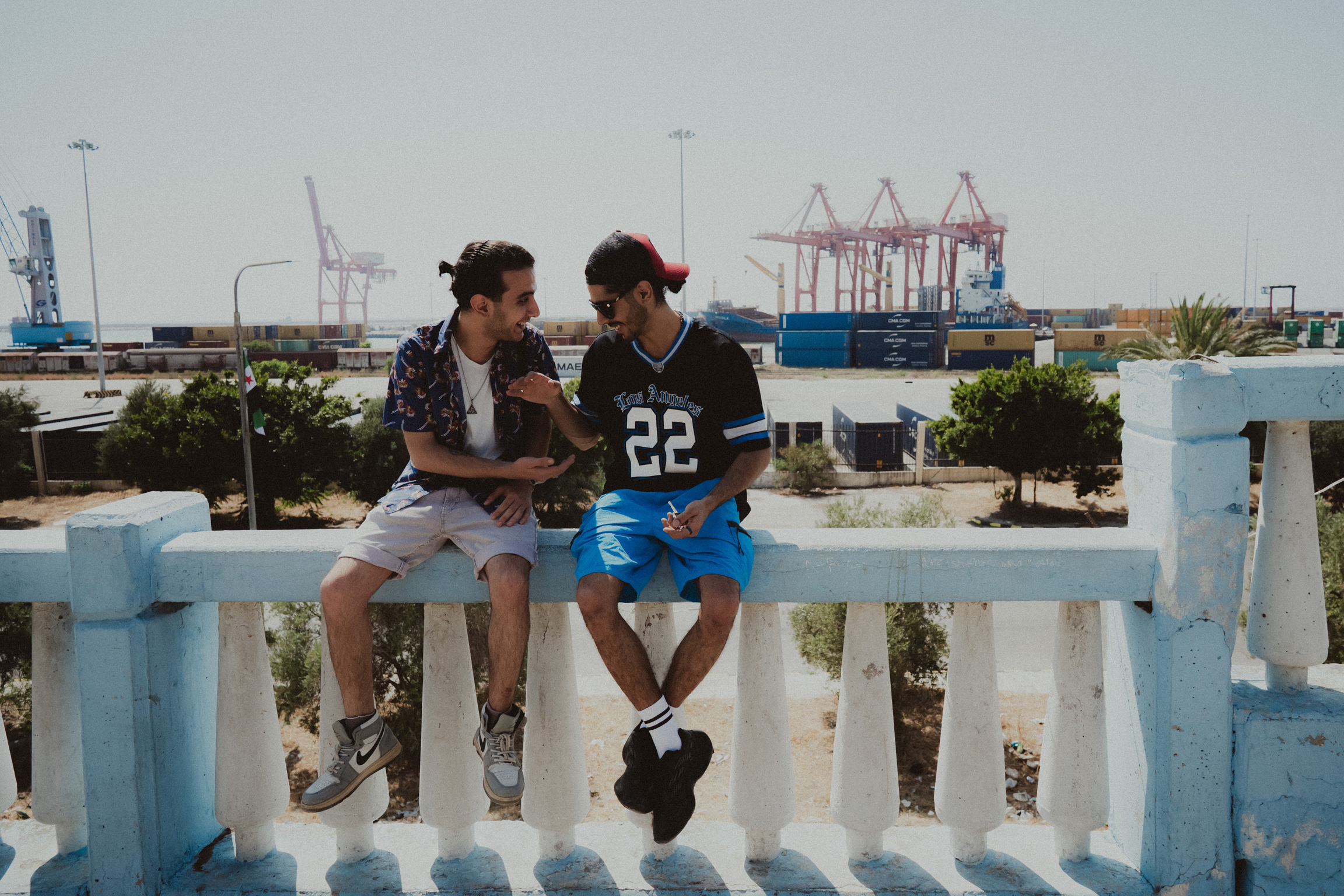Two young men sitting on white railing overlooking industrial port with cranes and containers in background.