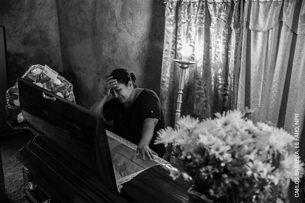 A black and white photo showing a person sitting on a chair, head in hands, with flowers and curtain visible in the background.