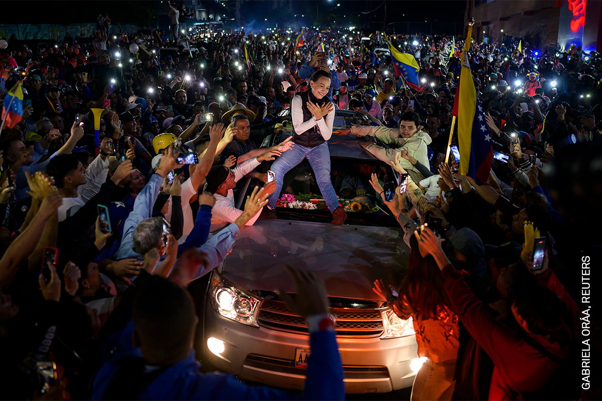 Nighttime protest scene, large crowd gathered, waving flags and holding lights, person standing on raised vehicle in centre.