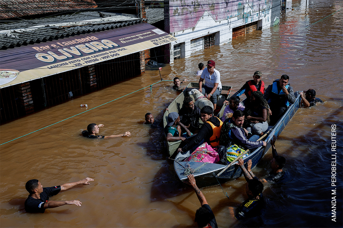 Flooded urban area with people wading through water, using boats to evacuate, on a submerged road with a large sign visible.