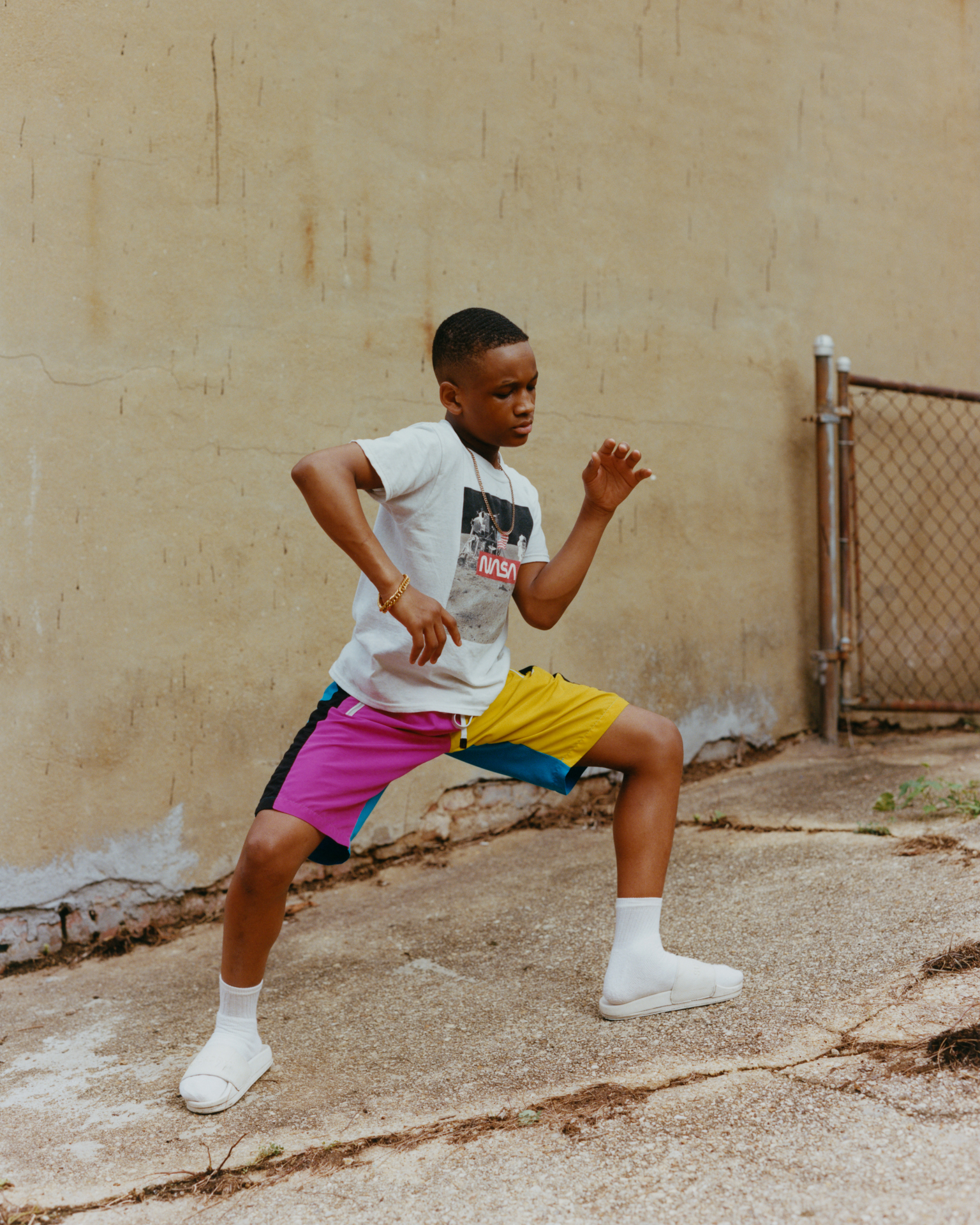 Young person dancing outdoors wearing white top and colourful purple-yellow shorts, against beige wall with chain-link fence.