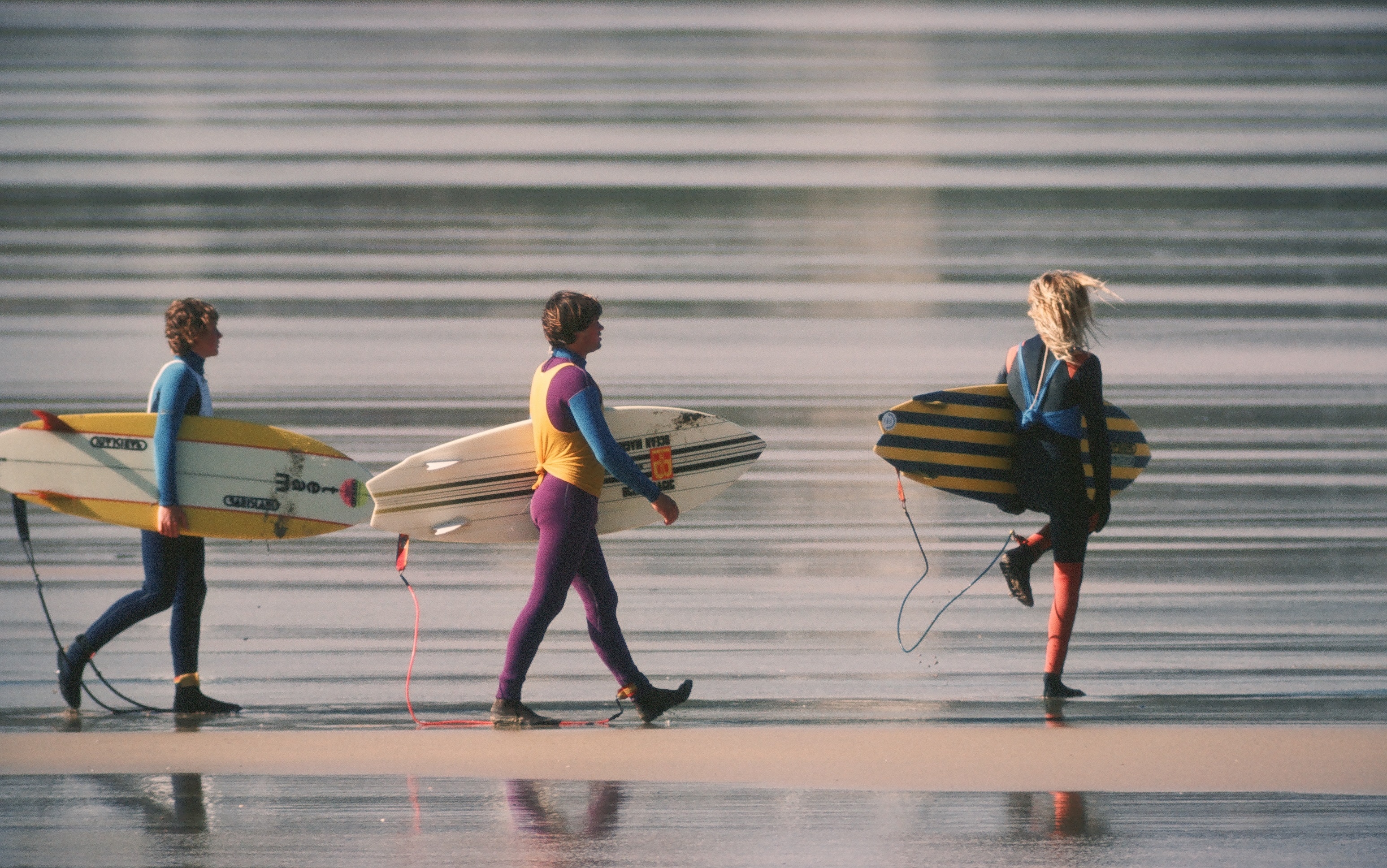 Three surfers walking on a beach, carrying their colourful surfboards.