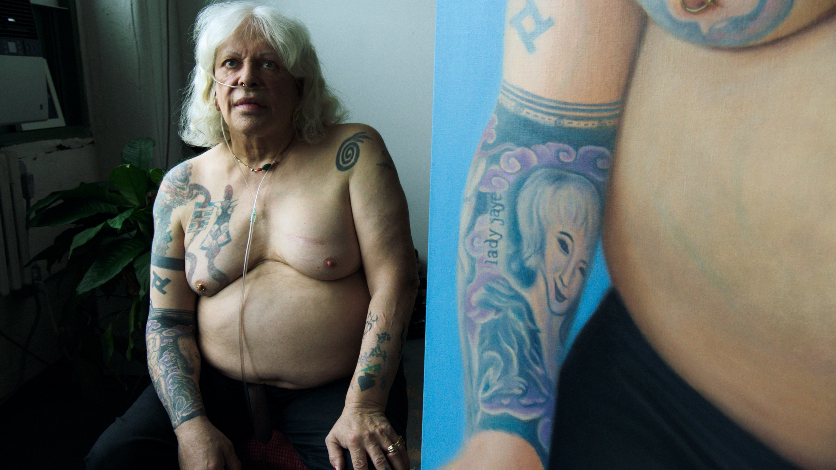 Elderly woman with white hair and extensive tattoos on arms and torso, seated shirtless indoors beside person with blue tattoos.