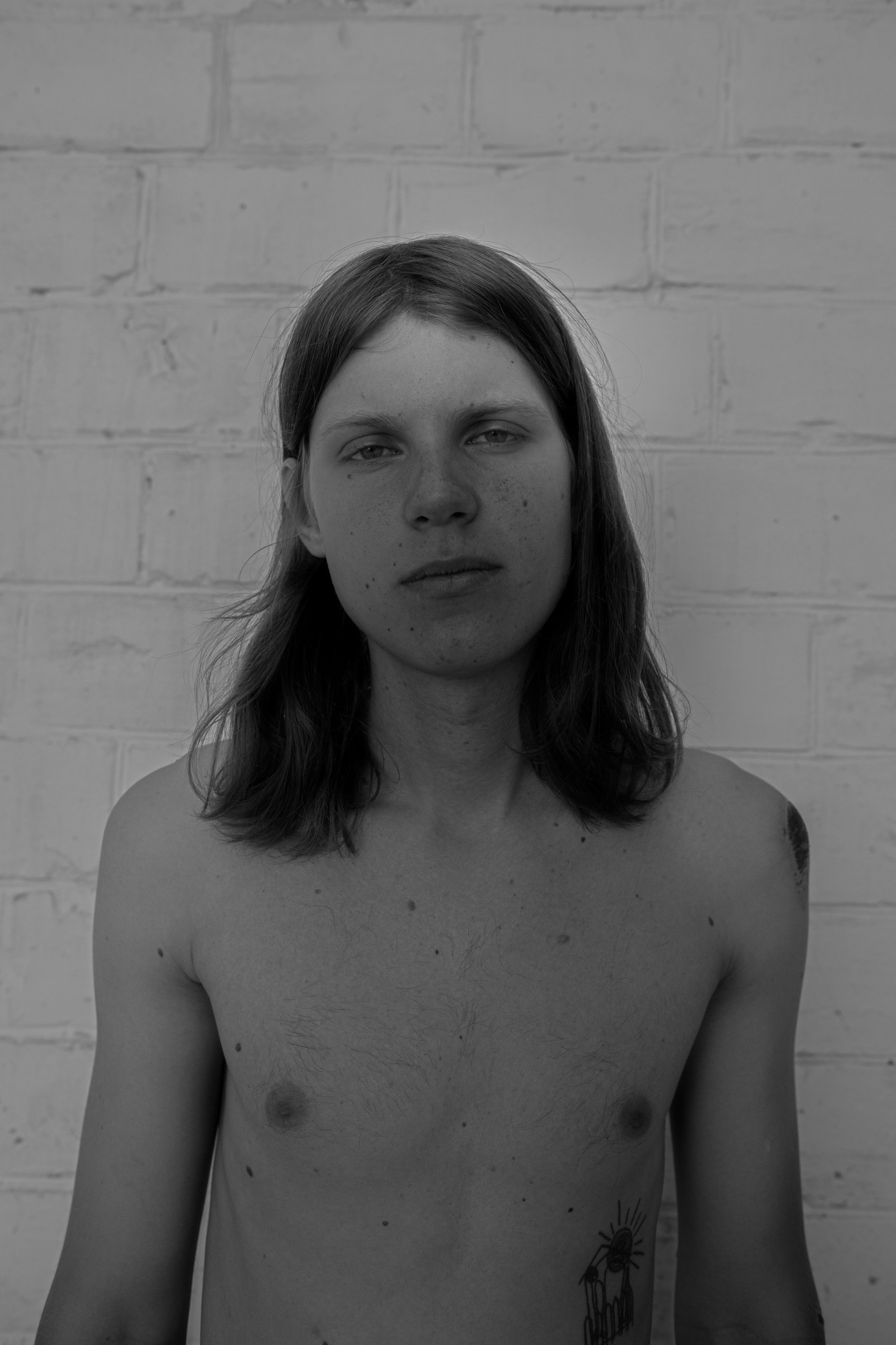A black and white portrait of a young person with shoulder-length hair against a white brick wall.