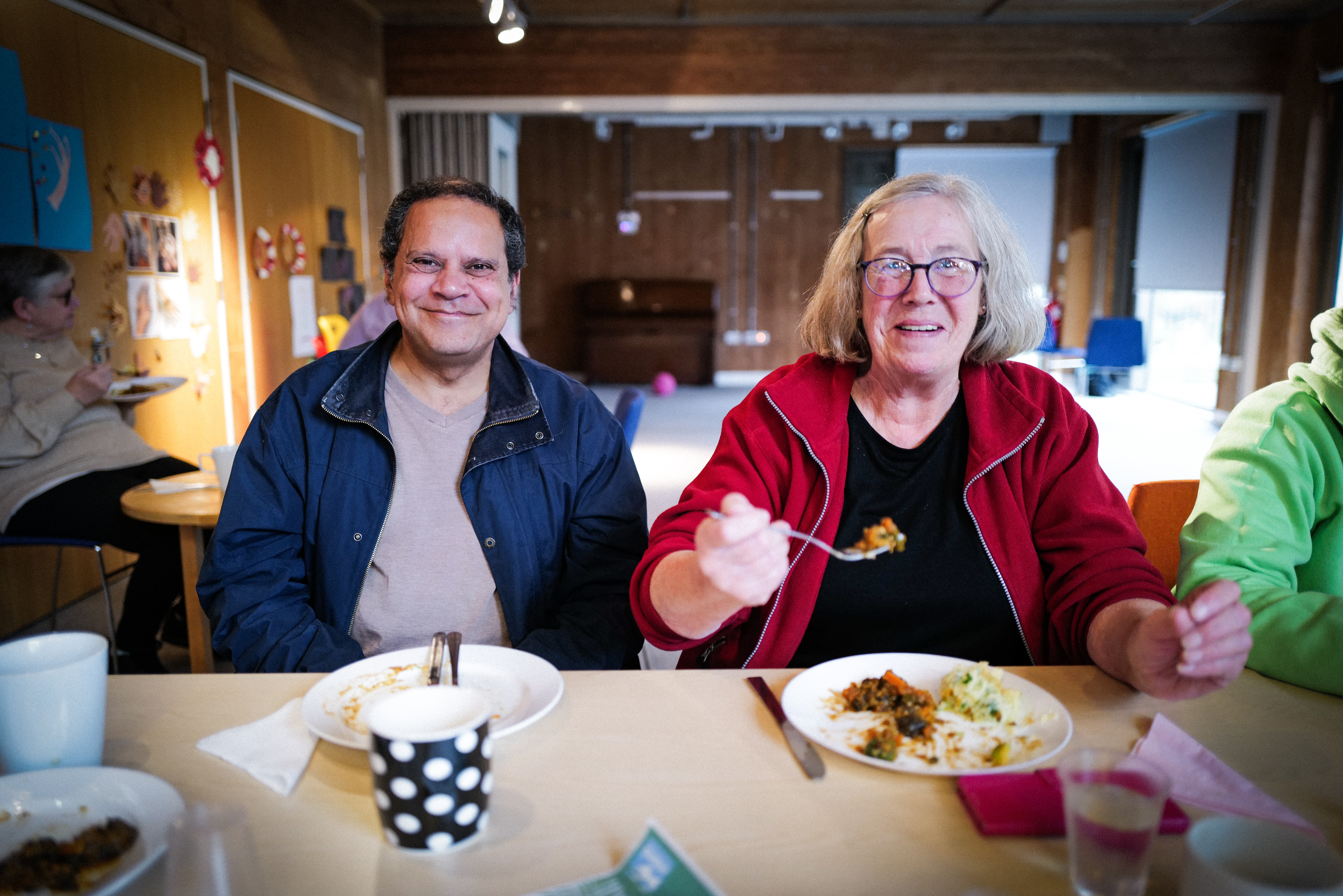 Two people sitting at table in community centre - man in dark jacket, woman in red cardigan with glasses, plates and polka dot mug visible.