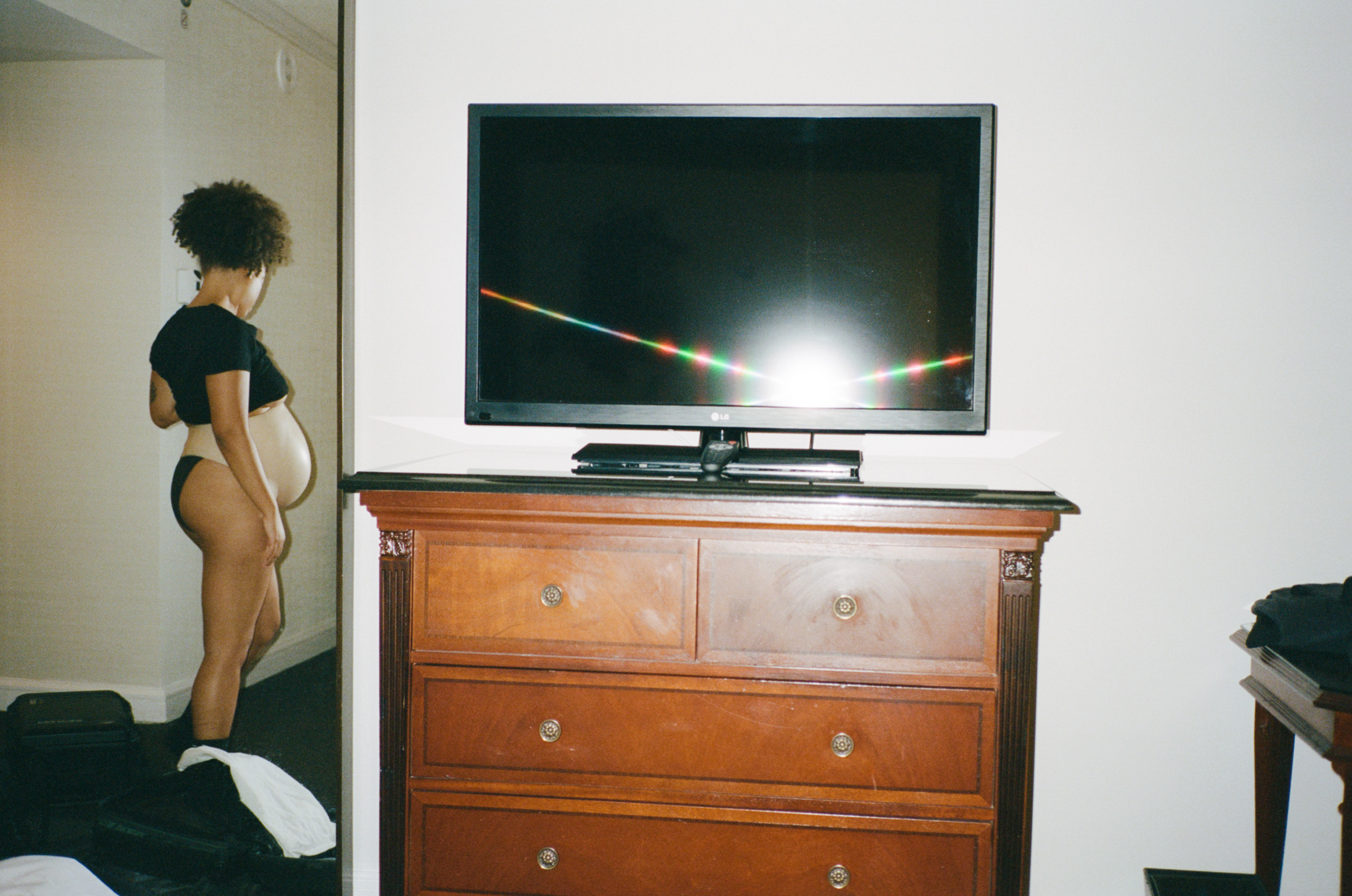 Woman in black clothing stands beside wooden chest of drawers with flat-screen TV on top in white-walled room.