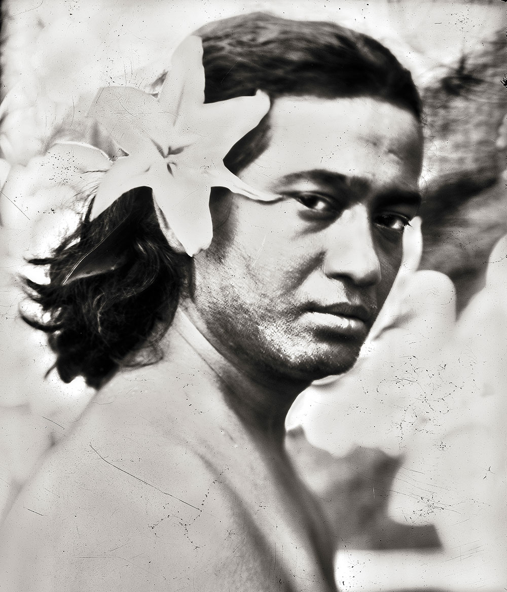 Black and white portrait of man with white plumeria flowers, one behind ear and one partially covering face, textured background.