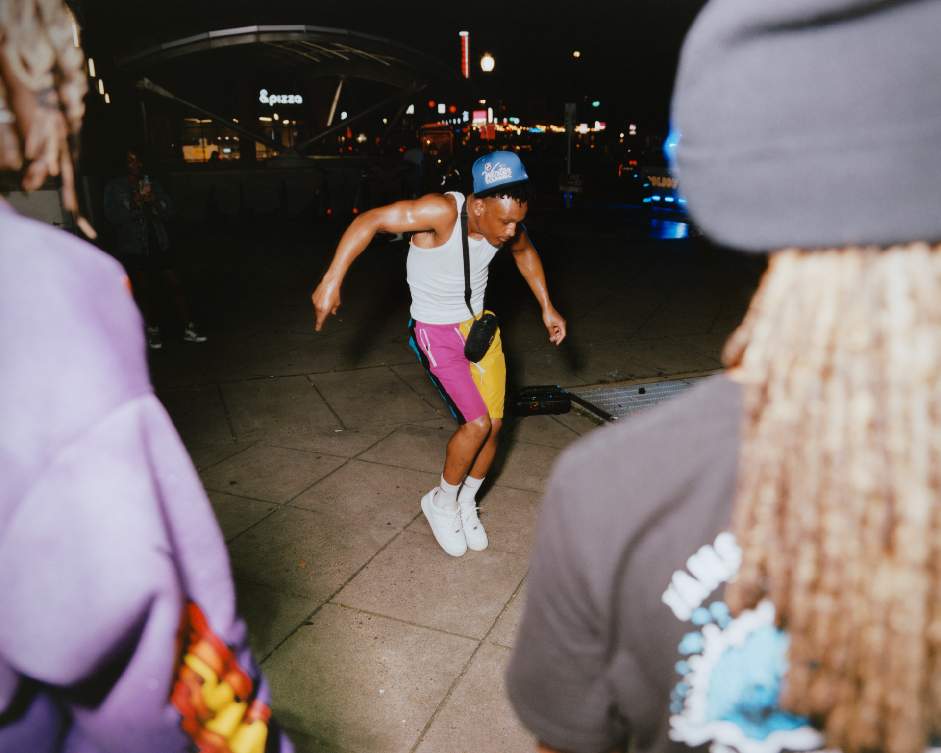 Person dancing in white top and pink leggings on tiled floor at night, surrounded by others in casual clothing.