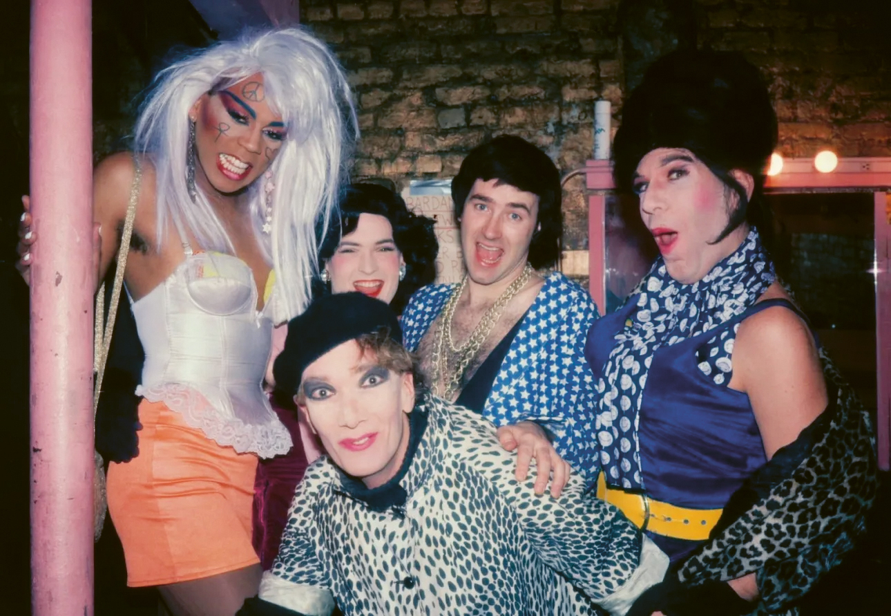 A group of five individuals in colourful, flamboyant clothing posing together in front of a dark background with pink accents.