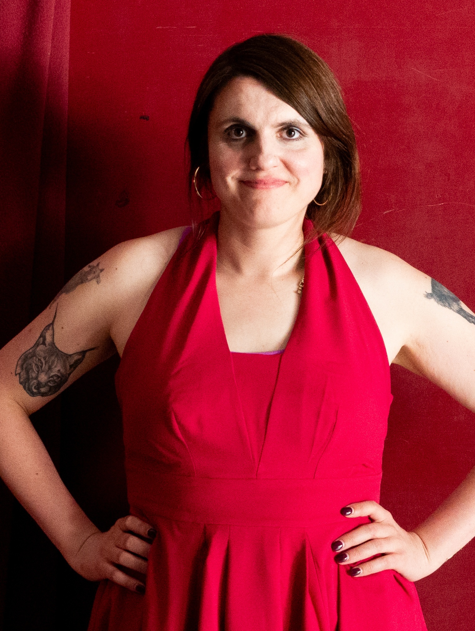 Woman with brown bob haircut wearing red halter dress, hands on hips, standing against red wall. Visible tattoos on both arms.