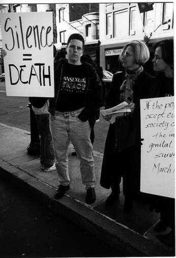 Black and white image showing protesters holding signs including "SILENCE = DEATH" and partial text on other placards, people standing on pavement.