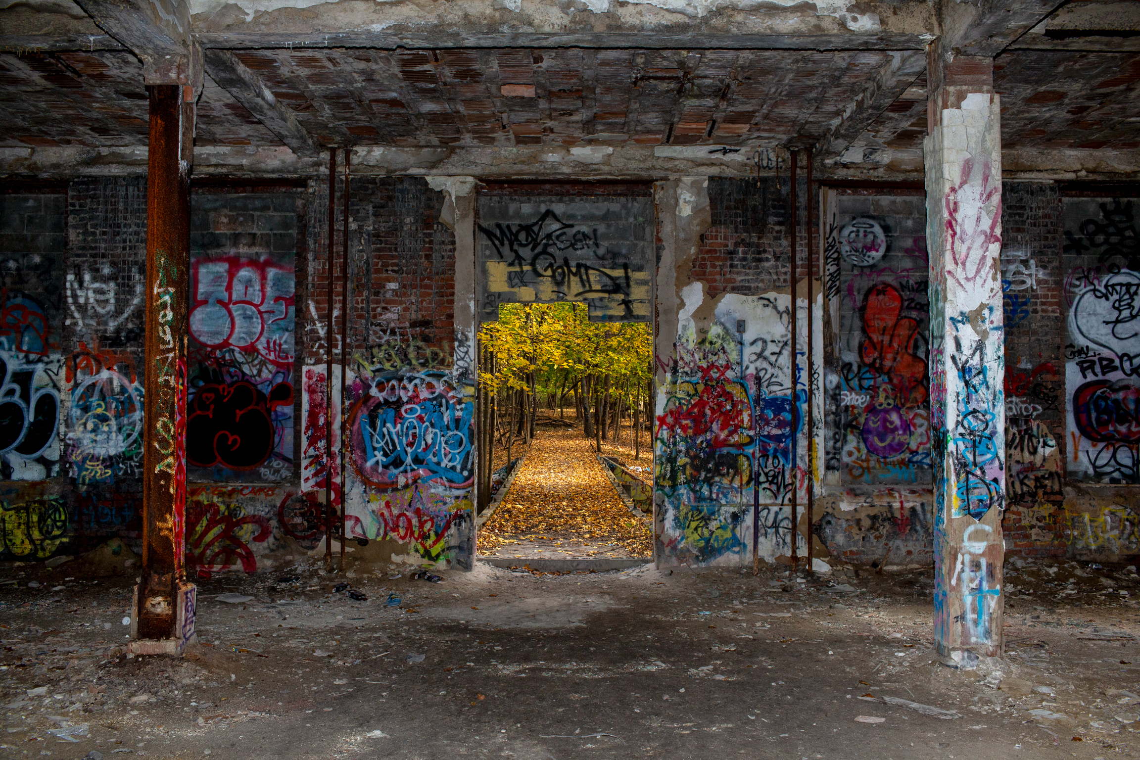 Derelict interior with concrete pillars and doorways covered in colourful graffiti, bright yellow light visible through far opening.