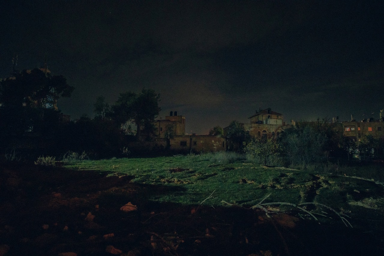 Dark night landscape with trees, buildings, and reflections in a body of water.