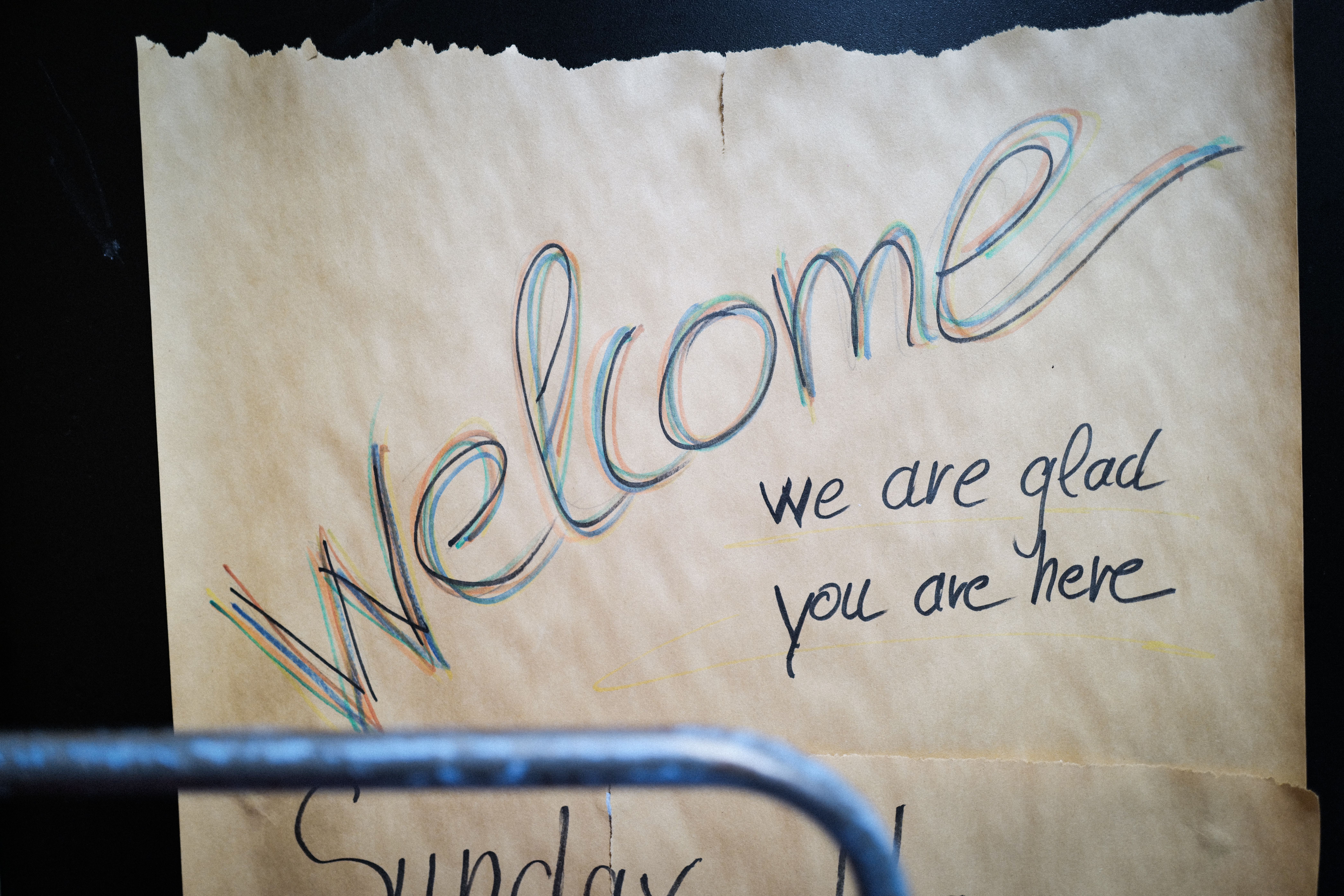 Handwritten note on cream paper with "Welcome" in multicoloured cursive letters and "we are glad you are here" in black text below.