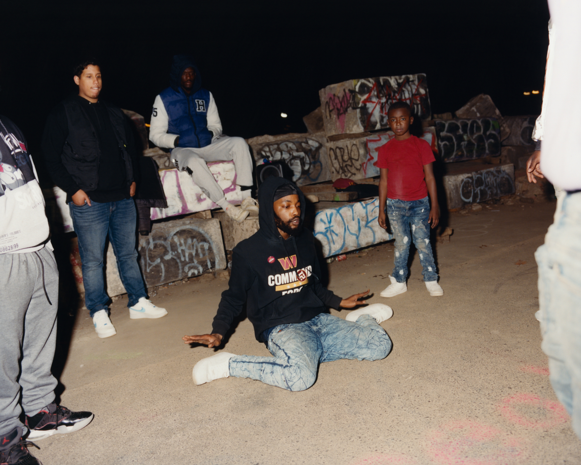Group of young people in underground concrete space with graffiti-covered walls, one person sitting cross-legged on floor.