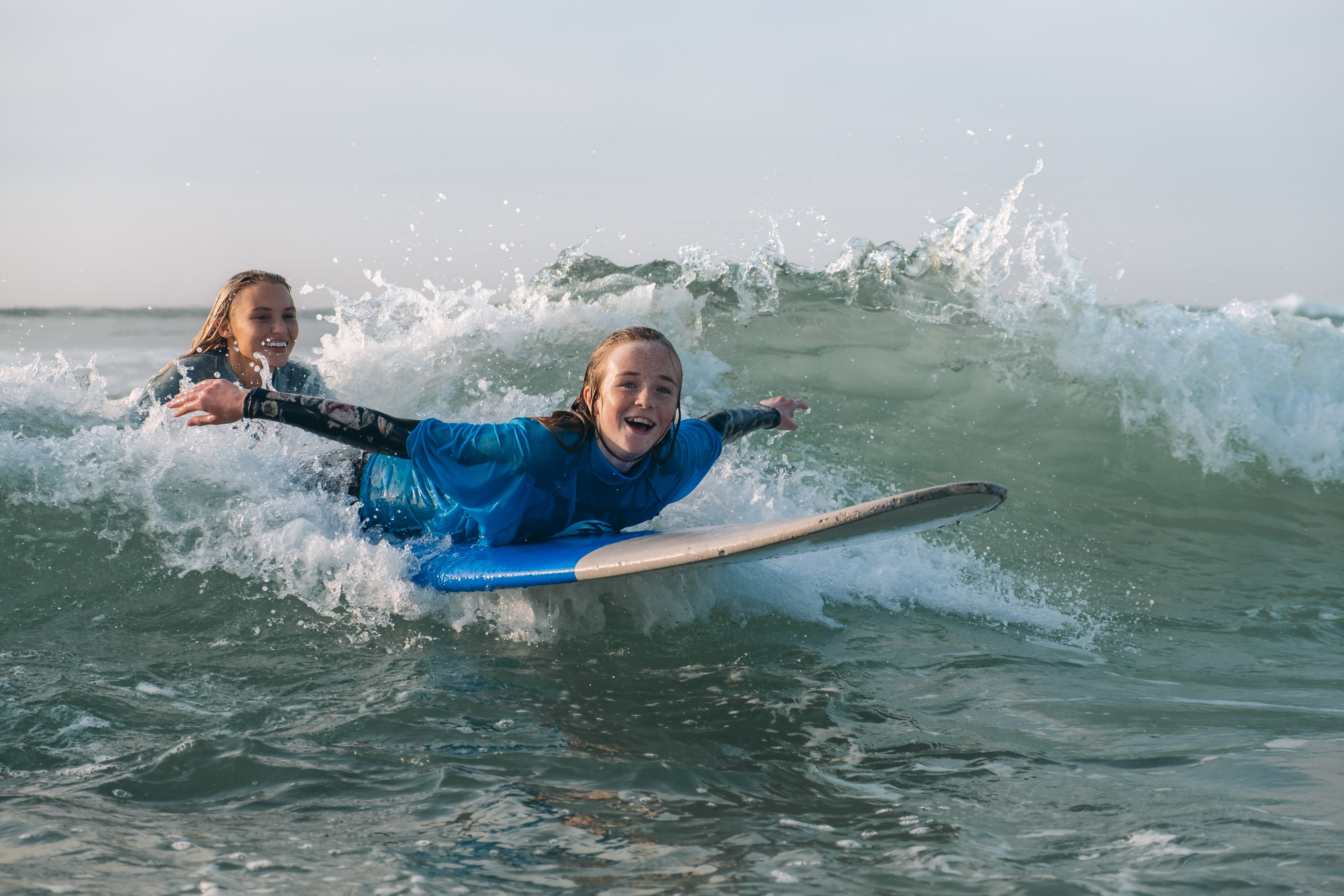 Two women surfing on waves, wearing blue wetsuits and riding surfboards.