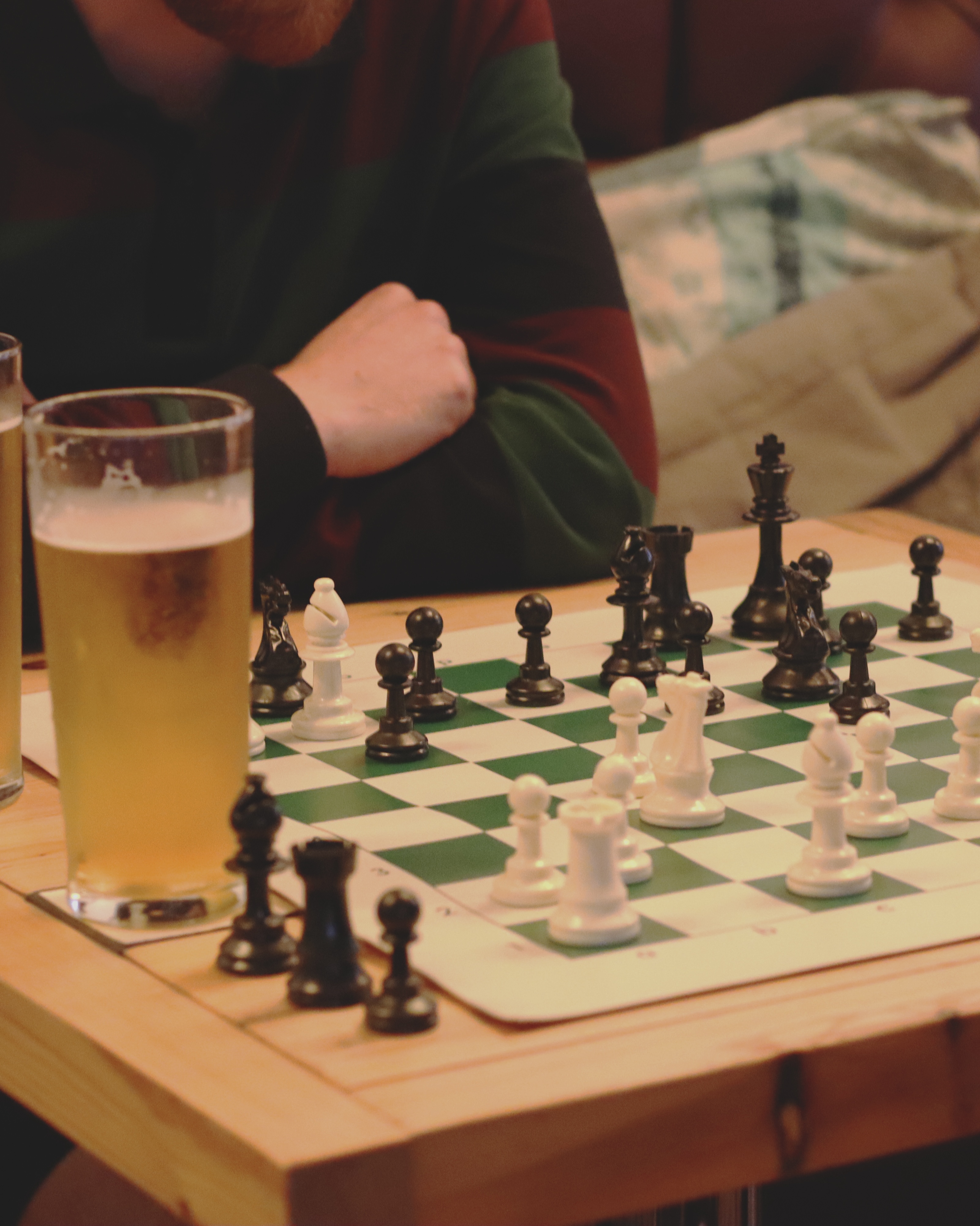 Chess set with black and white pieces on green and white chequered board beside glass of lager on wooden table.