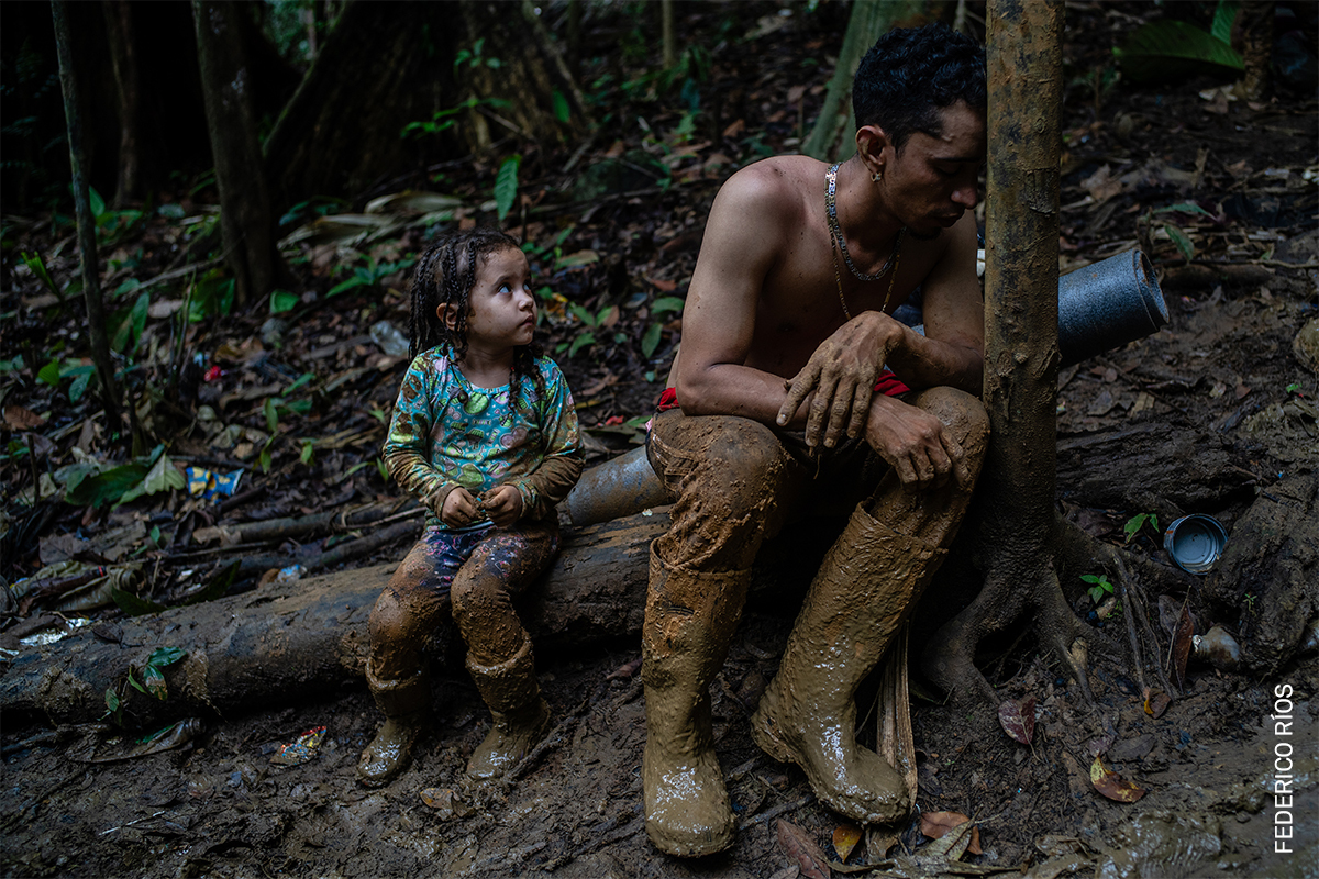 Two individuals, an adult male and a young child, seated on a fallen tree trunk in a lush, green forest environment.