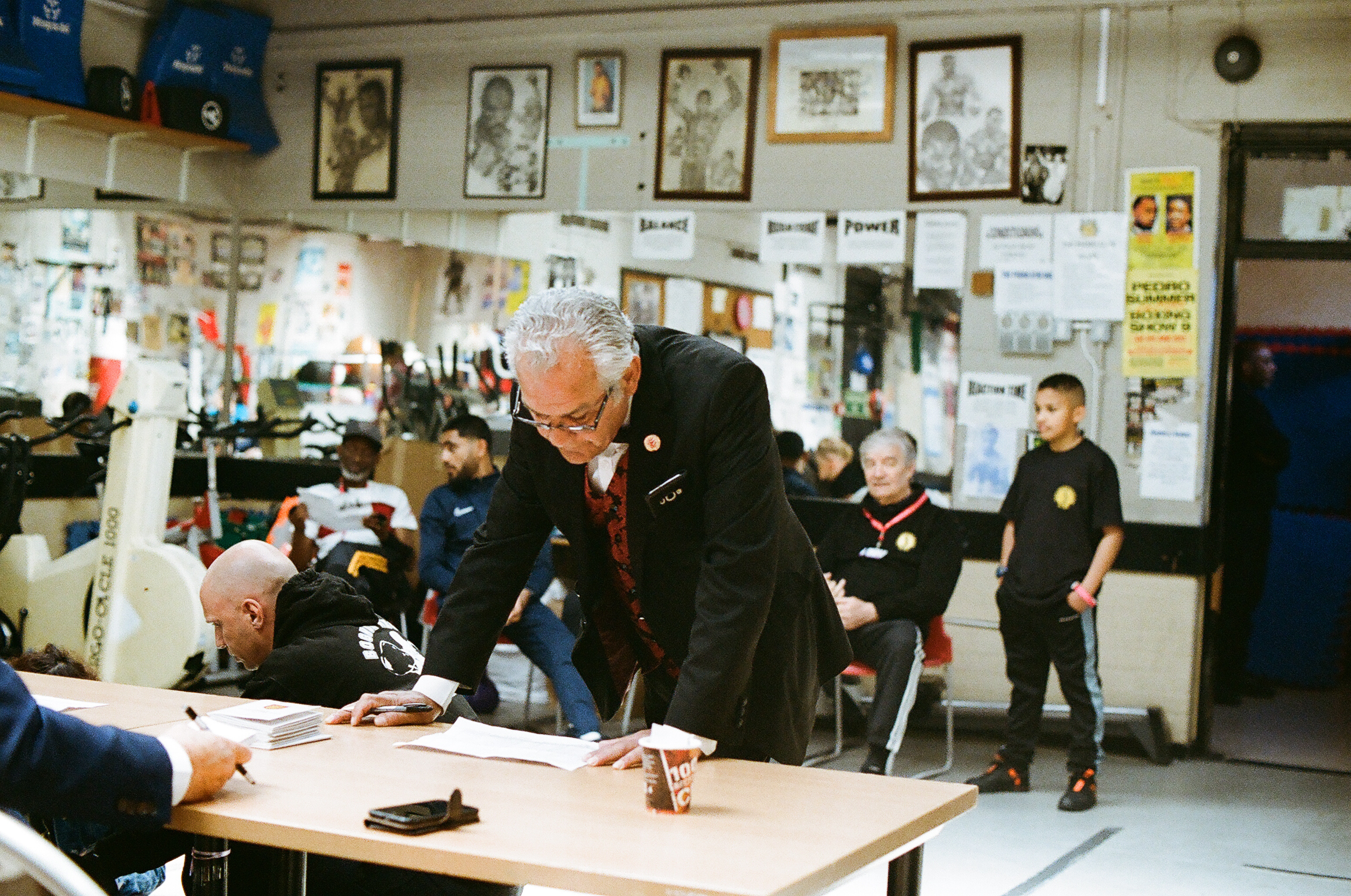 Elderly man in dark uniform leans over table with papers whilst others sit in background. Wall displays framed photographs and documents.